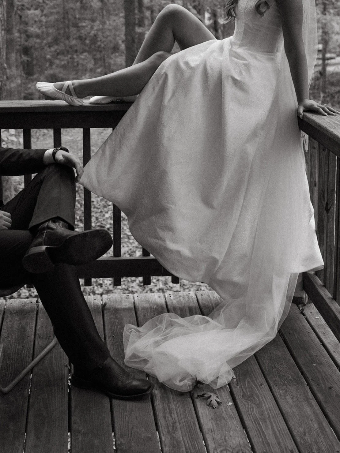 Close-up of a woman in a wedding dress sitting on a porch with one leg crossed and wearing ballet shoes, while a man in a suit sits nearby with his leg stretched out.