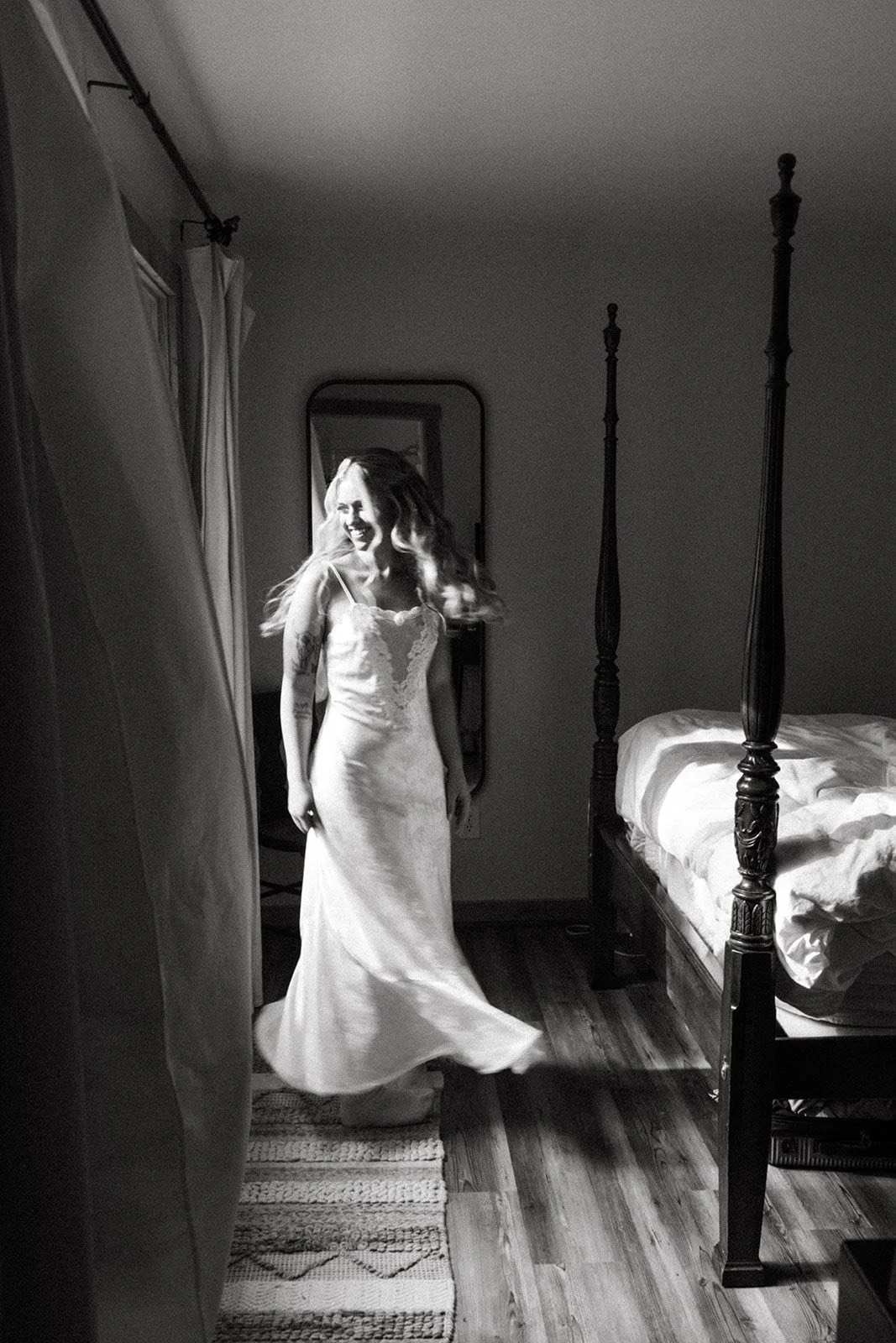 A woman in a wedding dress smiling and twirling in a bedroom with a four-poster bed, mirror, and curtains.