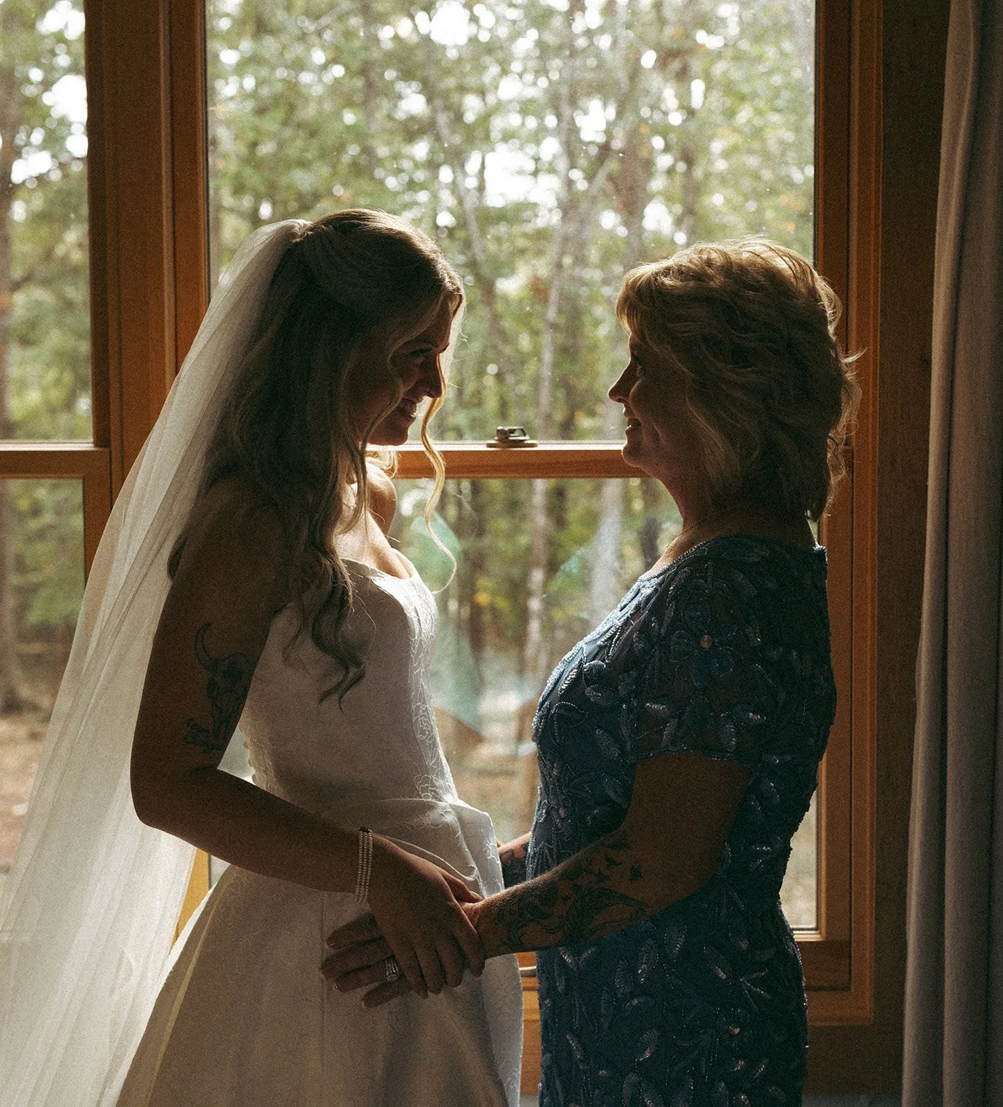 A bride in a wedding dress and veil holding hands with a woman in a floral dress, standing by a window with trees outside, shadowed inside.