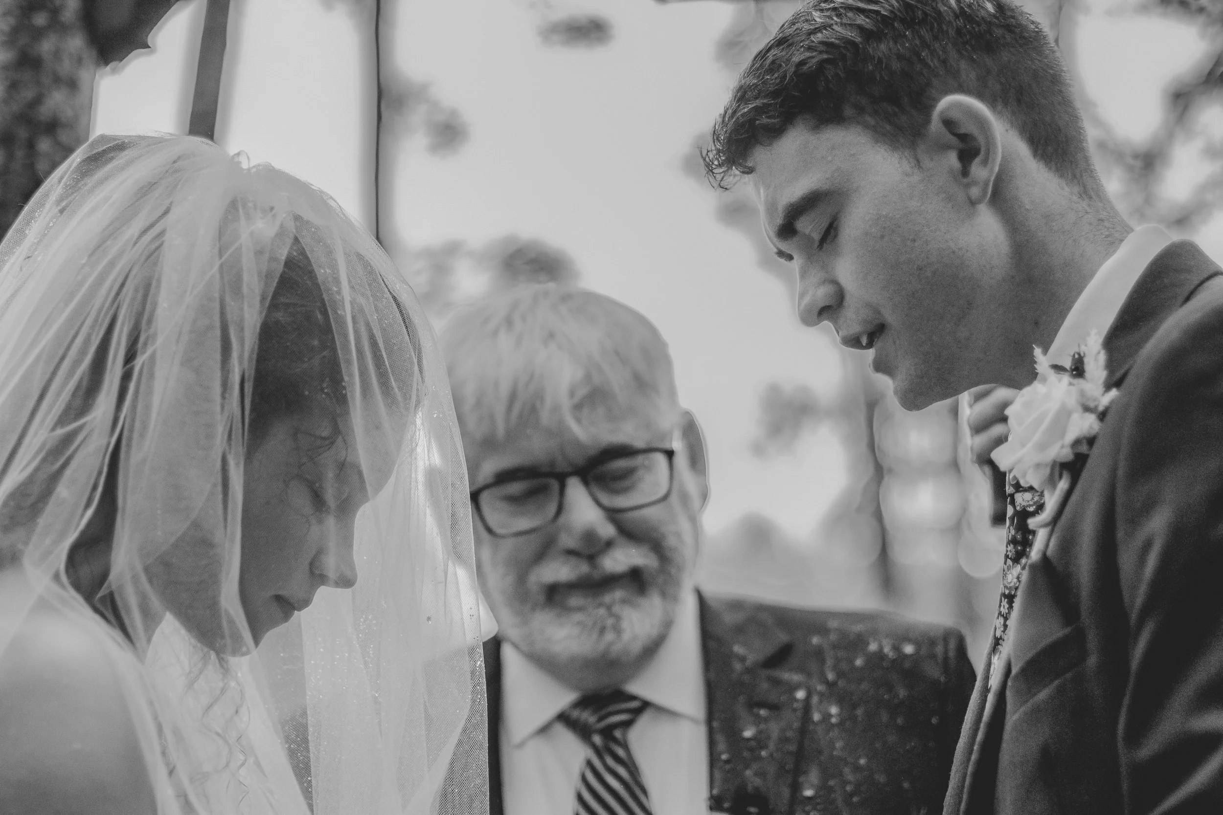Black and white photo of a wedding ceremony showing a bride with a veil, a groom wearing a suit with a boutonniere, and a celebrant or minister with glasses and a striped tie, all close together with heads bowed.