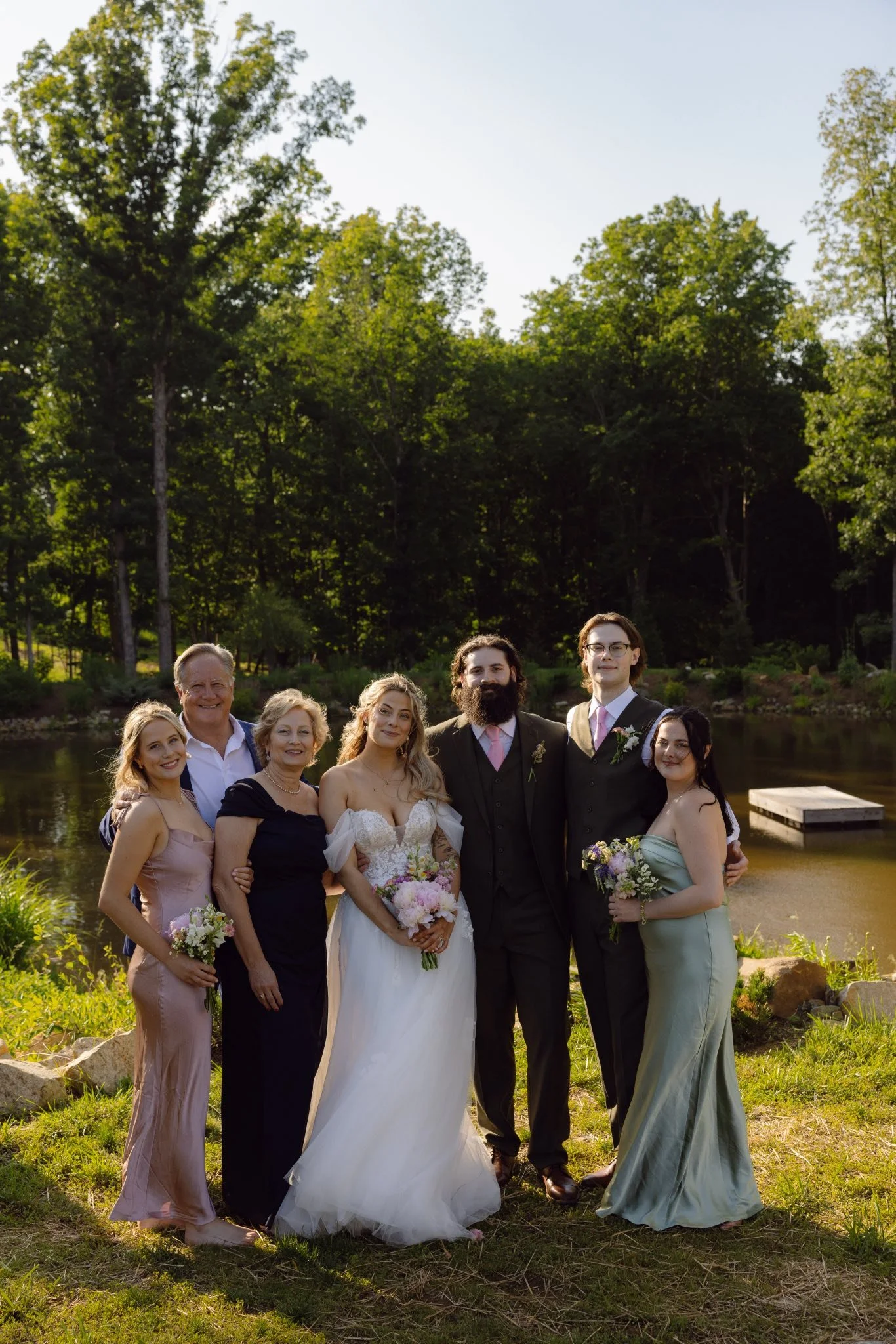 Group of people at a wedding outdoors by a lake with green trees in the background.