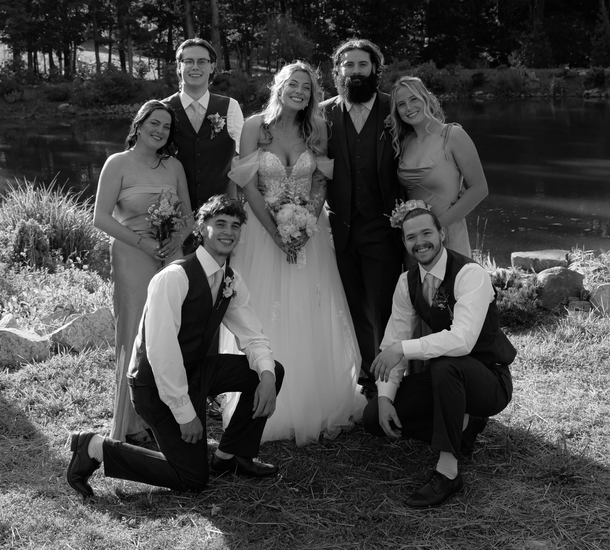 A group of nine people at a wedding outdoors near a pond, including the bride in a wedding dress holding a bouquet, groom, bridesmaids, groomsmen, and other guests, all smiling.