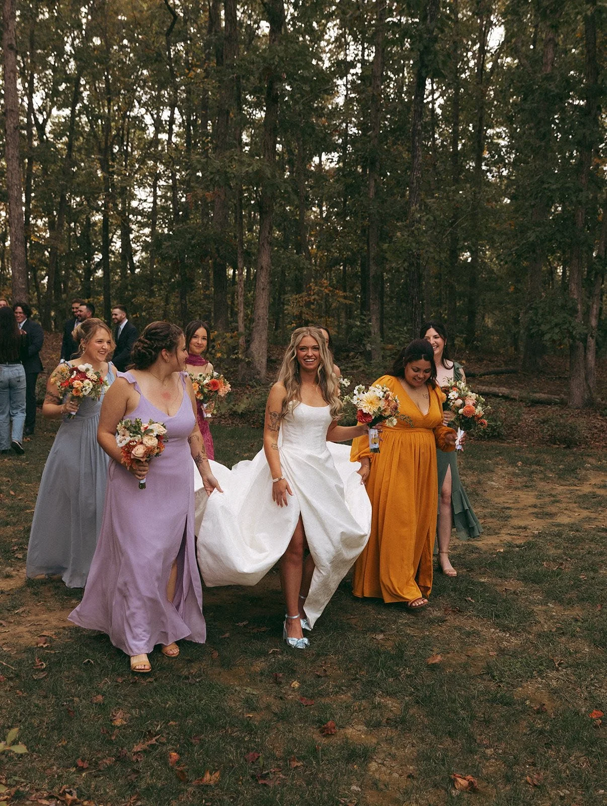 Women dressed in colorful dresses walking outdoors in a wooded area, holding bouquets of flowers, during a wedding celebration.
