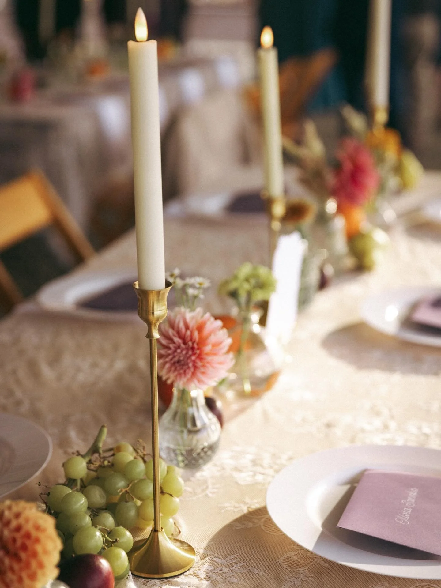 Elegant event table setup with tall white candles in gold holders, pink and orange flowers in glass vases, grapes, and plates with pink napkins.
