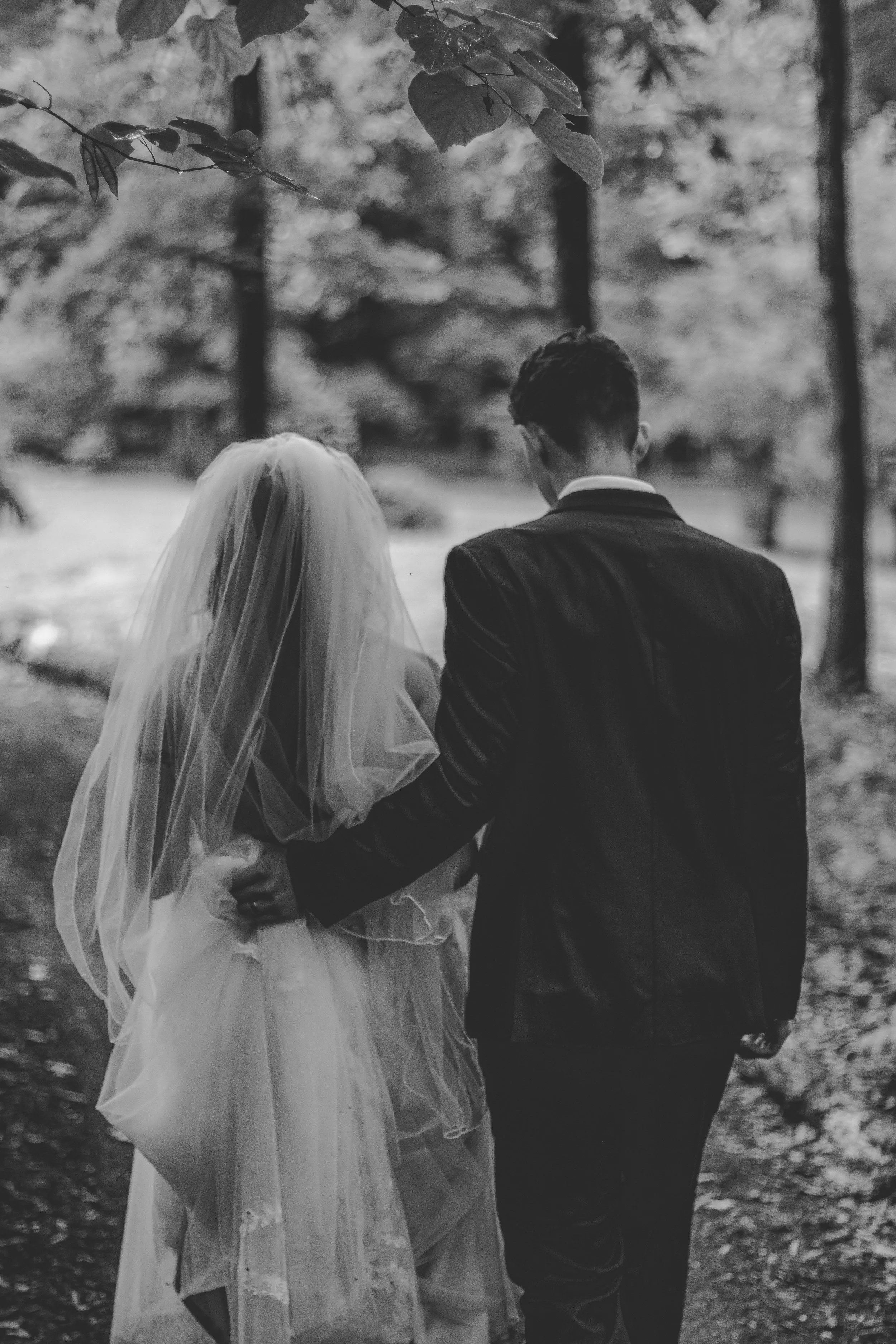 A black-and-white photo of a bride in a wedding dress and veil holding hands with a groom in a dark suit walking outdoors among trees.