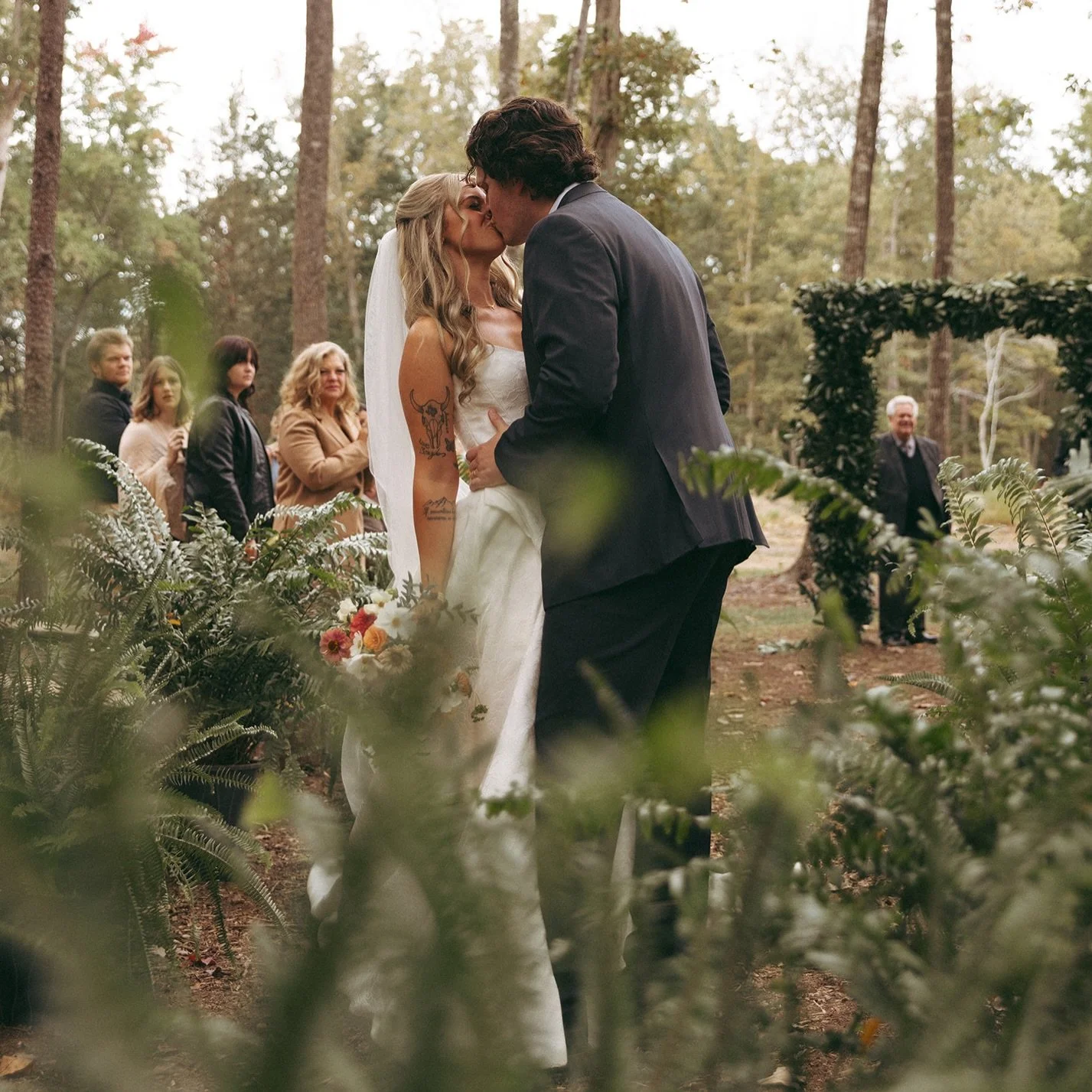 A couple shares a kiss during a wedding ceremony in a wooded outdoor setting, with bridal party members and guests observing in the background.