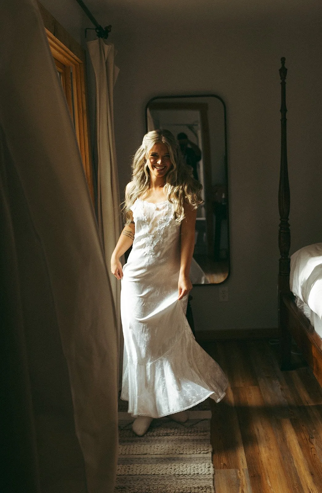 A woman in a white wedding dress standing in a dimly lit room, smiling and holding the bottom of her dress, with a mirror and a bed nearby.