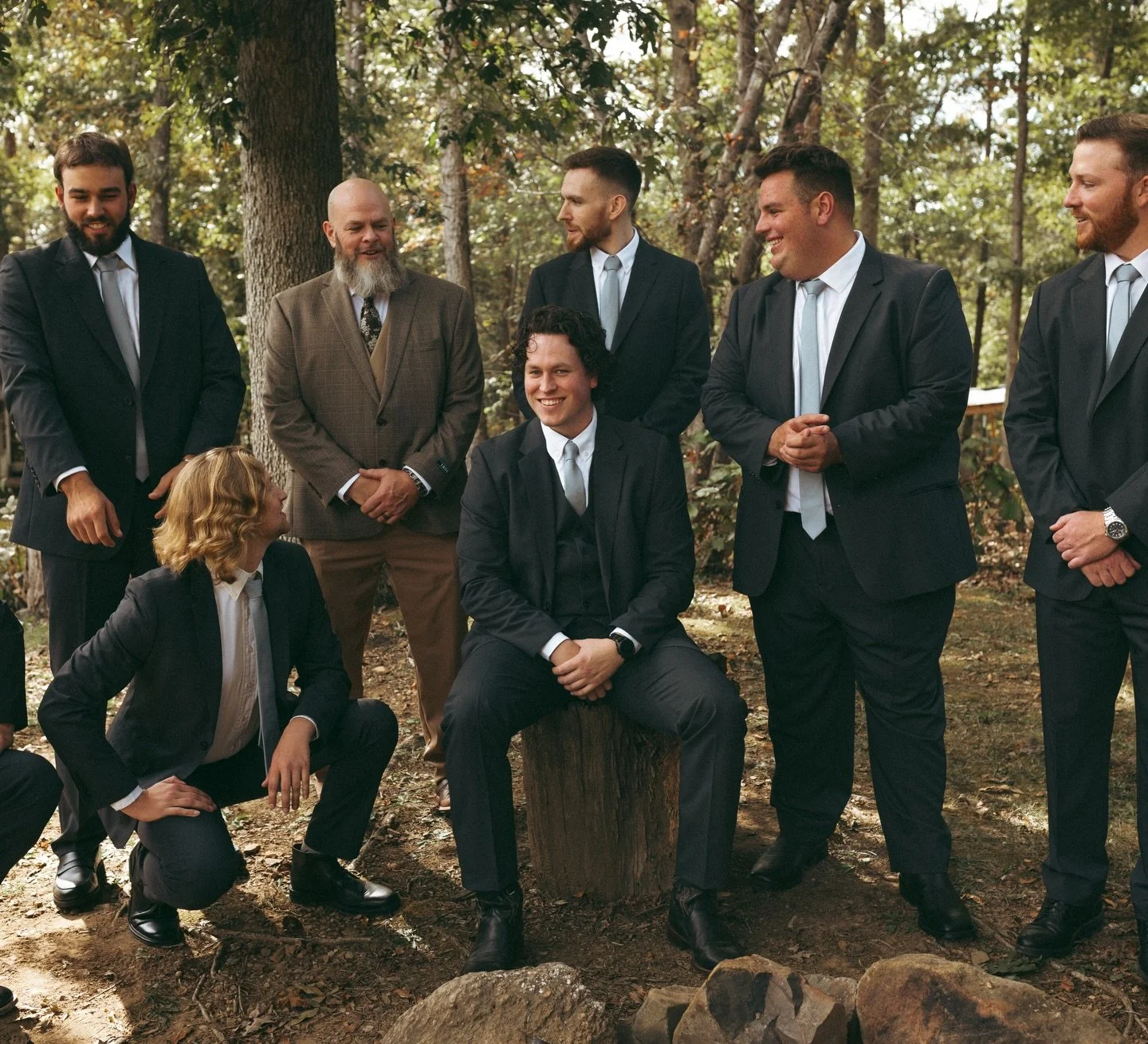 Group of men dressed in suits gathering outdoors in a wooded area, with some standing and one sitting on a tree stump, smiling and interacting.