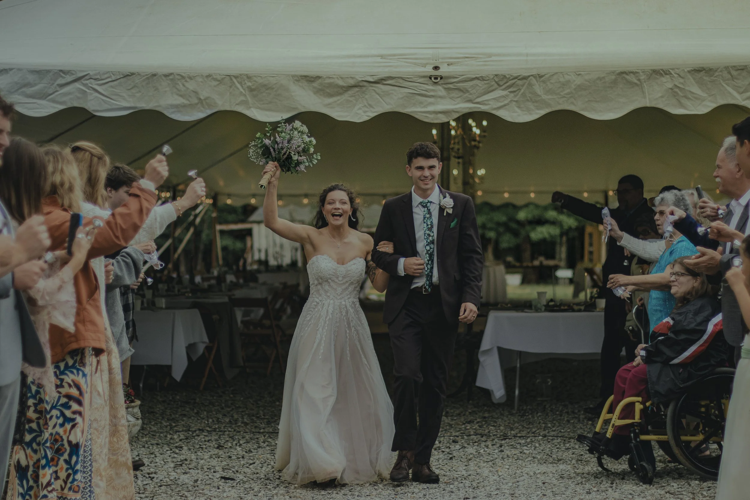 Bride and groom smiling and walking down the aisle at their wedding reception, surrounded by guests cheering and taking photos under a large outdoor tent.