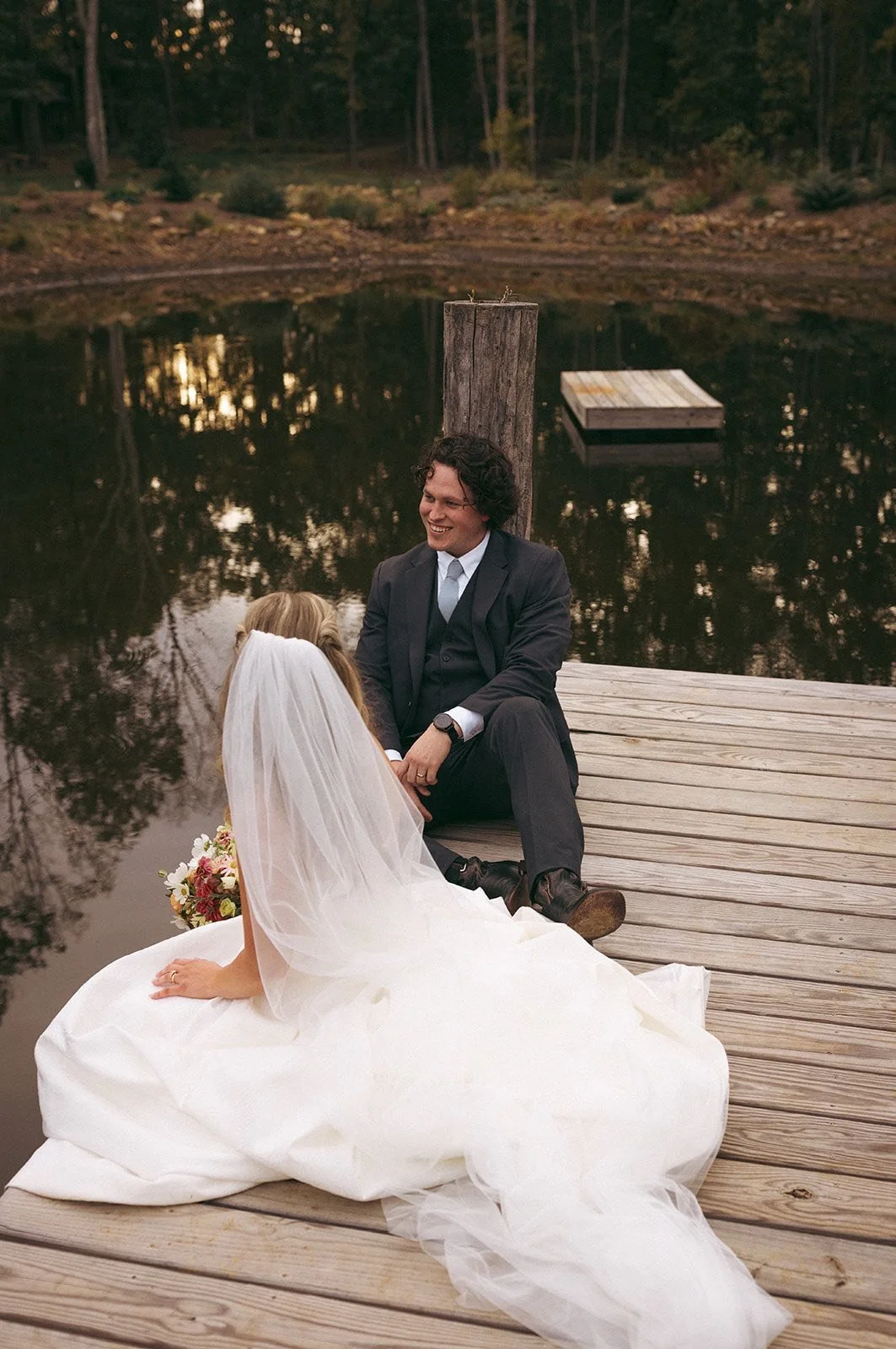 A bride in a white wedding dress and veil sitting on a wooden dock by a pond, talking with a man in a dark suit sitting near her.