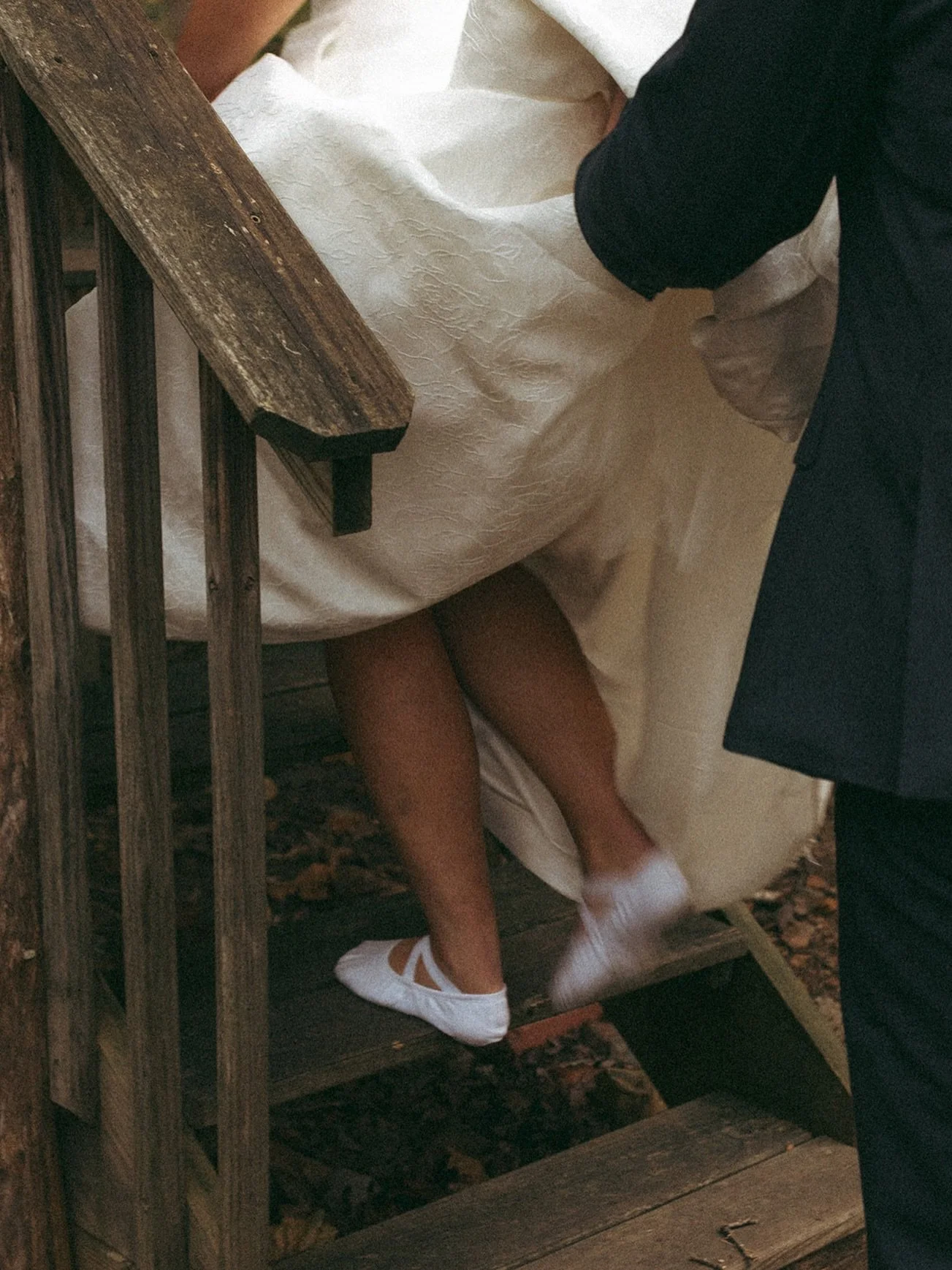 A person in a white dress is getting baptized in an outdoor setting, with a wooden platform and stairs, by a man in dark clothing.