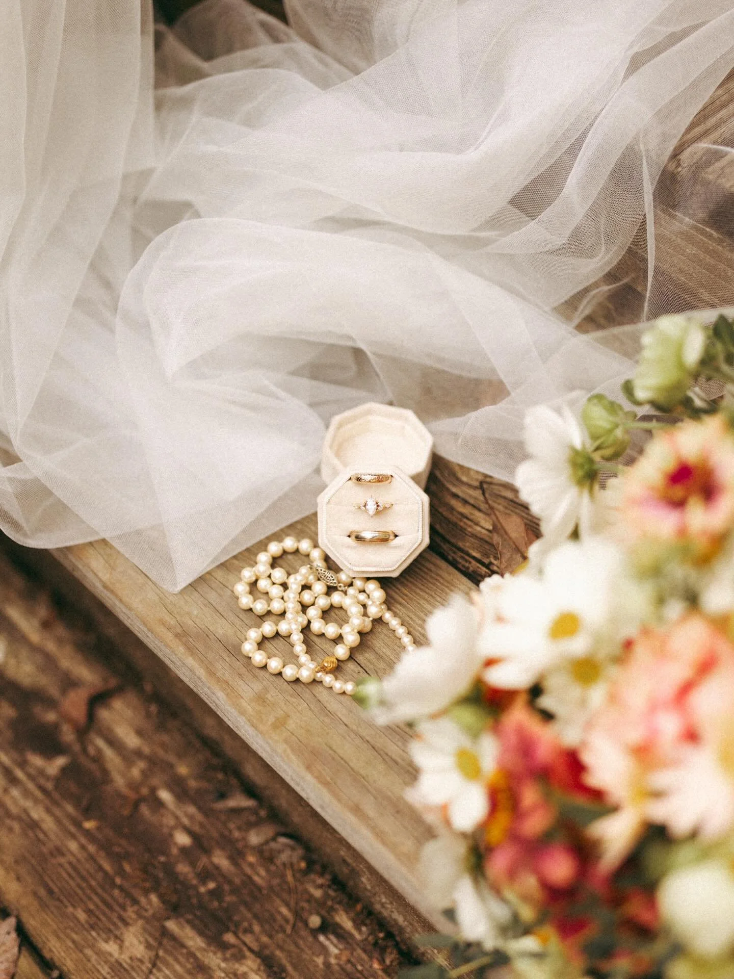 Wedding jewelry including pearl necklaces, wedding rings, and a diamond engagement ring in a velvet jewelry box, with a sheer veil, a wooden surface, and a blurred bouquet of flowers in the foreground.