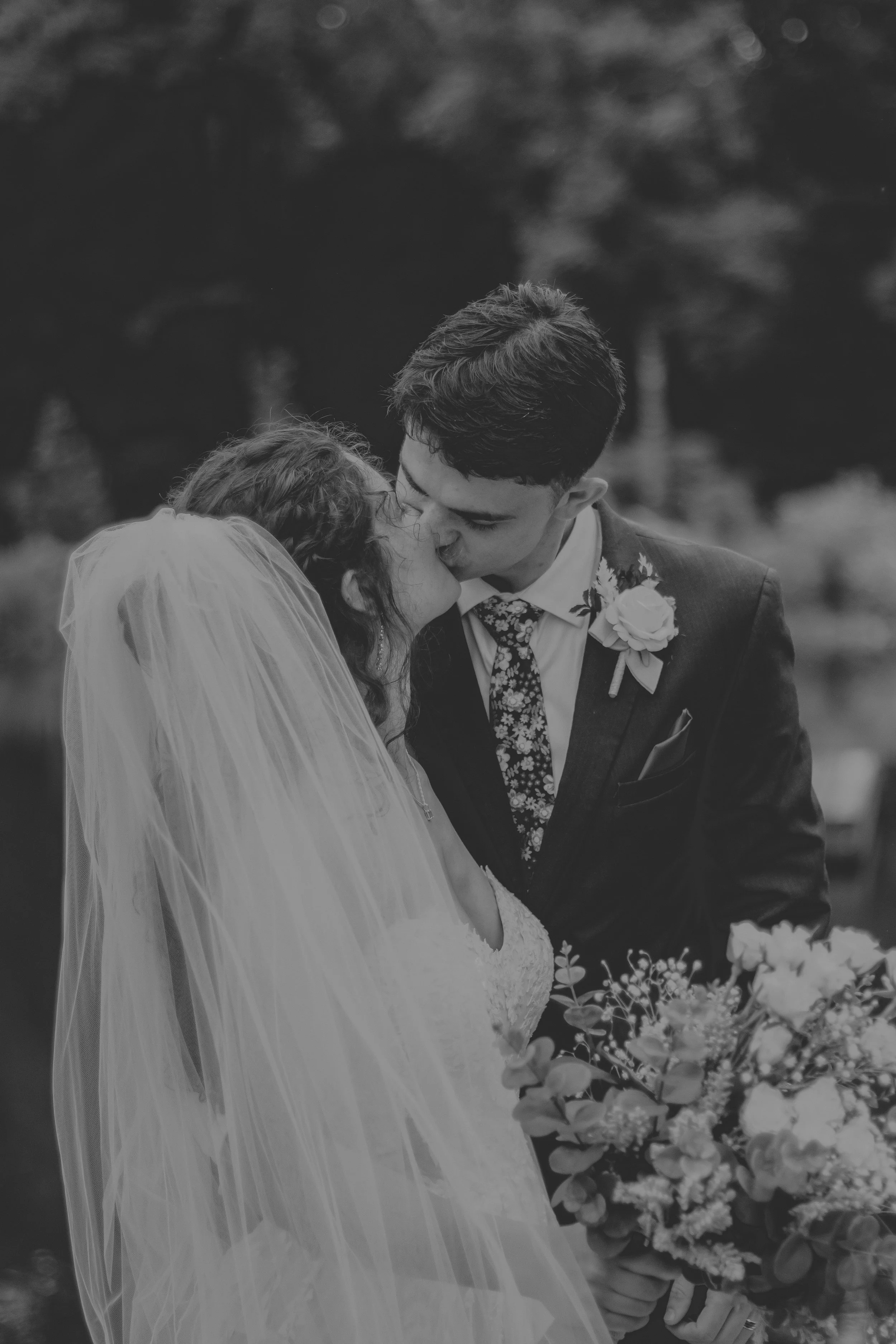 A bride and groom sharing a kiss during their wedding ceremony outdoors, with the groom holding a bouquet of flowers and wearing a suit with a floral tie and boutonniere.