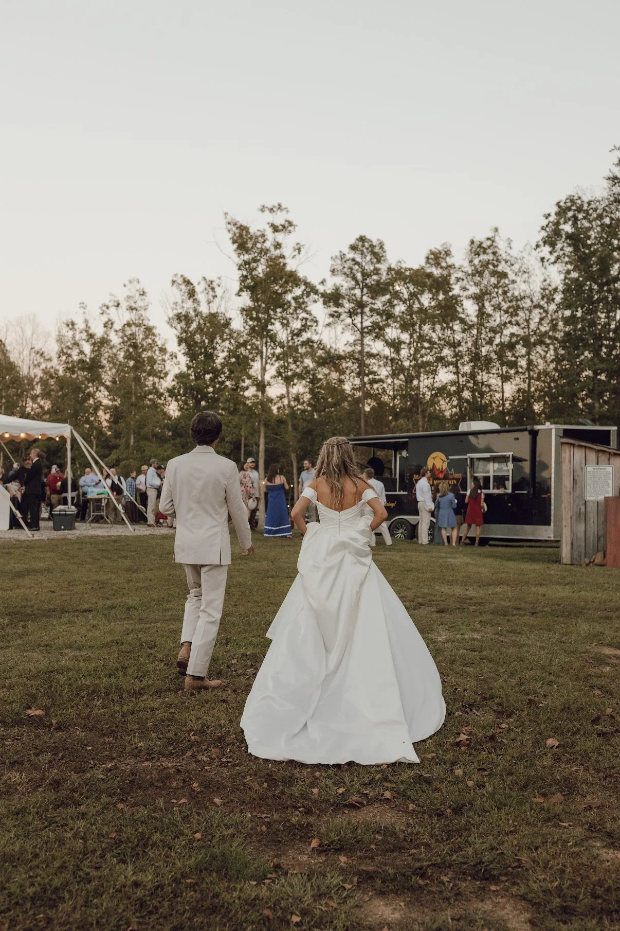 A bride and groom walking on grass towards a reception area with people, trees, and a food truck in the background at sunset.