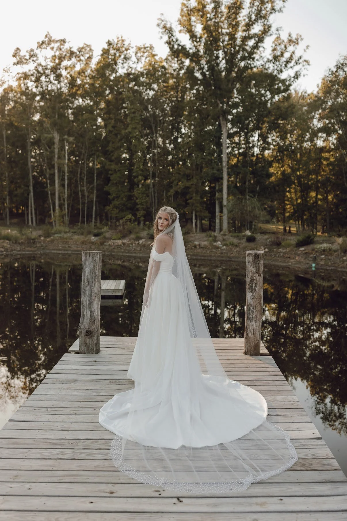 A bride in a white wedding dress and veil stands on a wooden dock by a calm lake at sunset, surrounded by trees.