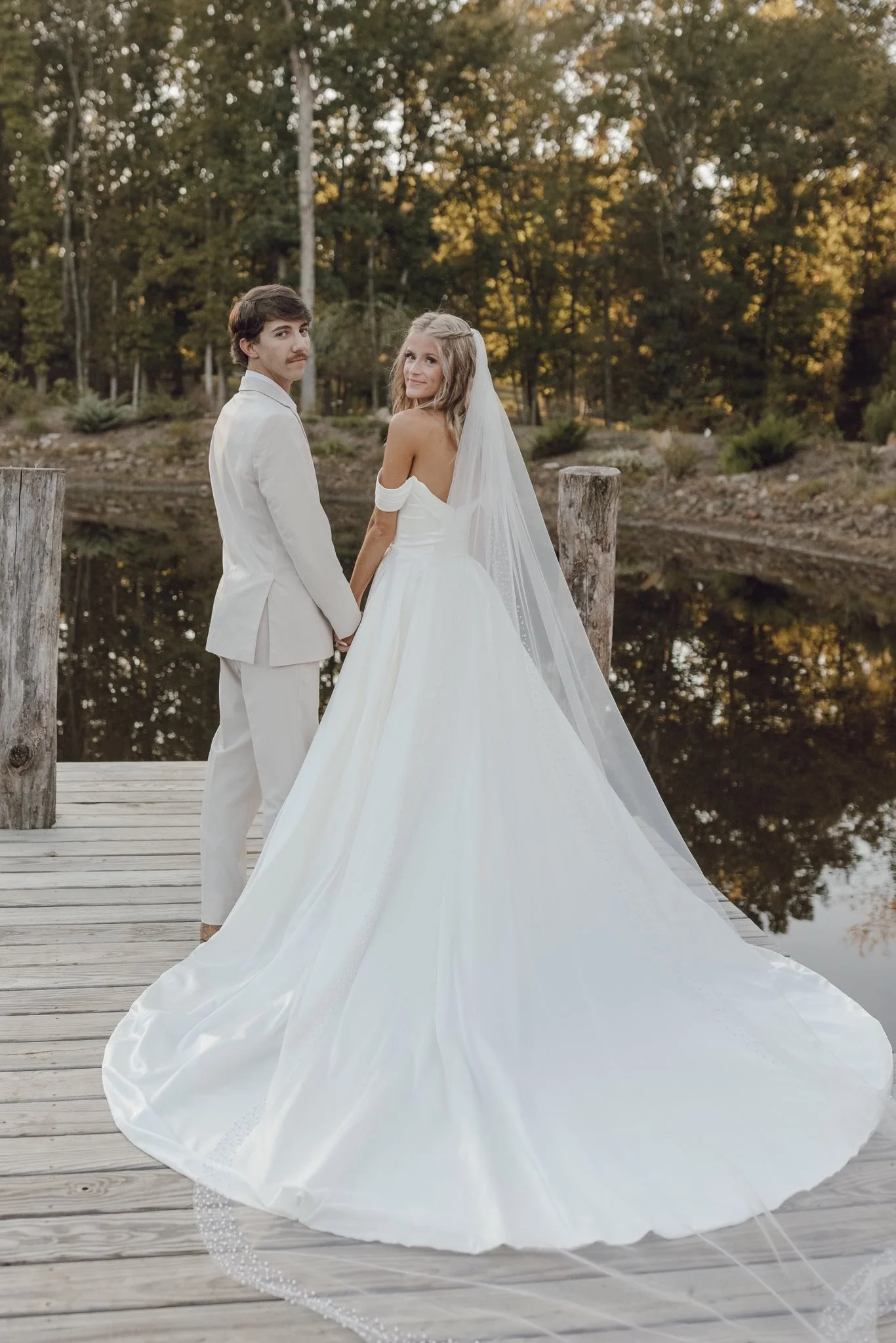 A bride and groom standing hand in hand on a wooden dock by a lake, surrounded by trees at sunset.