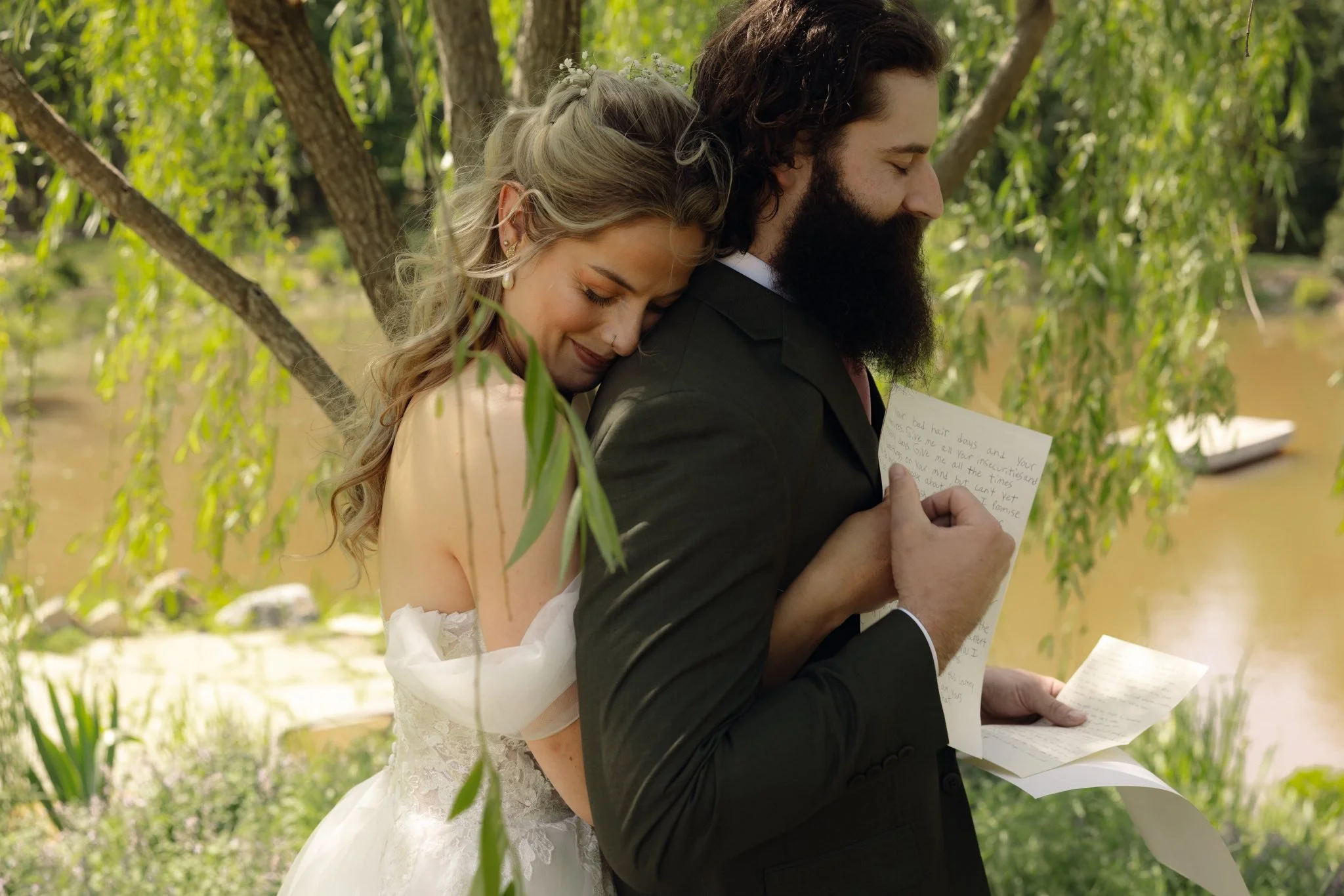 A bride and groom are reading heartfelt notes outdoors near a lake, with trees and greenery in the background.
