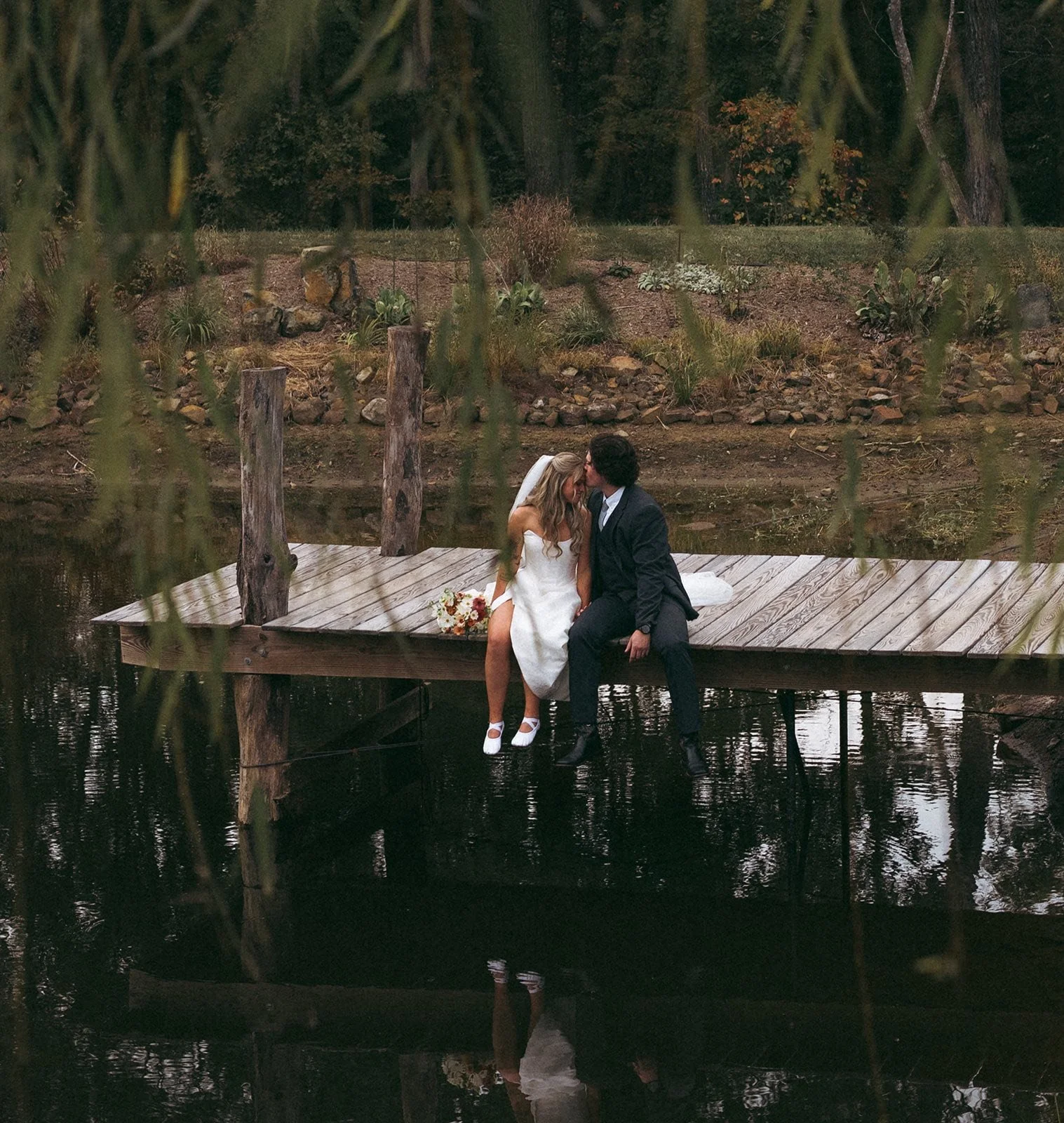 A bride and groom sitting on a wooden dock by a lake, sharing a kiss during their wedding, with a bouquet beside them and trees in the background.