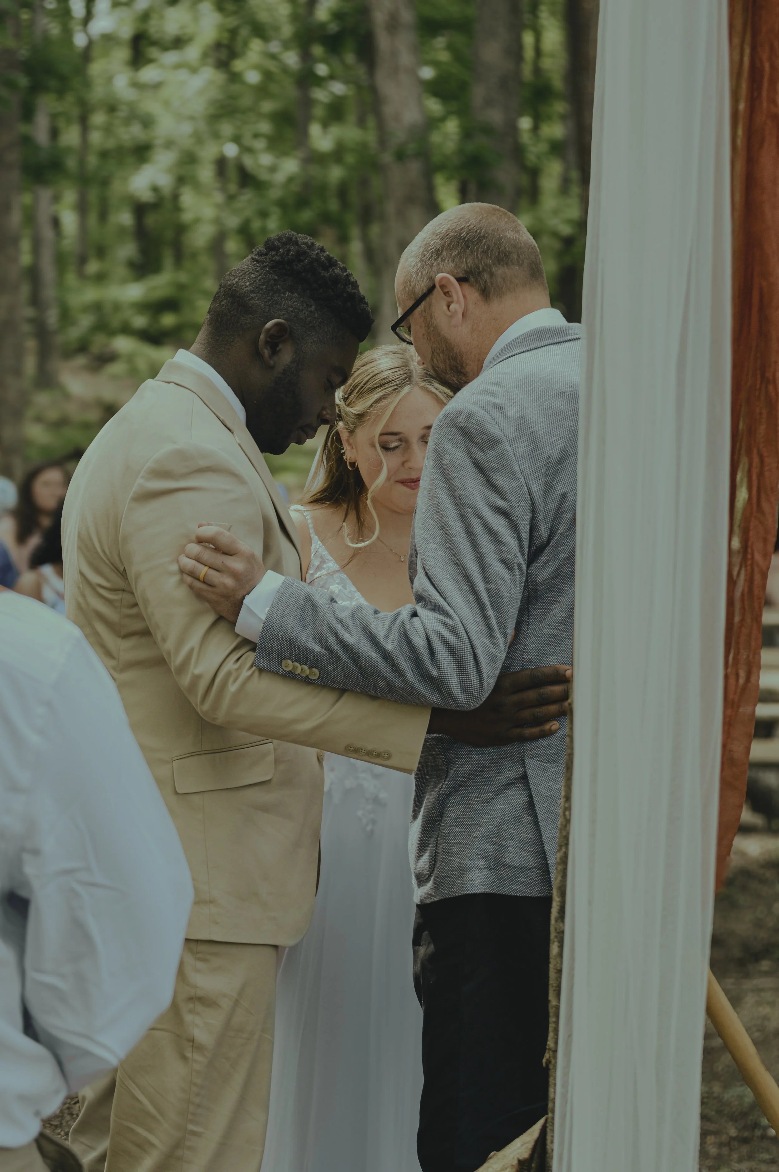 A couple getting married during an outdoor wedding ceremony with a person officiating, all standing close together with their eyes closed, surrounded by trees and greenery.