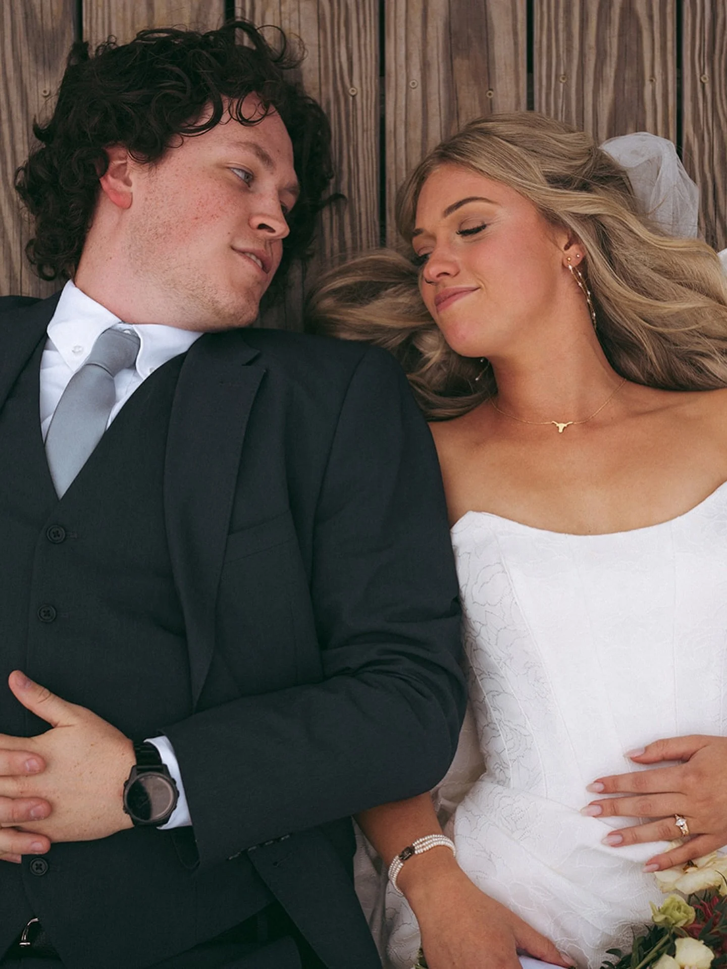 A newlywed couple resting on a wooden surface, smiling at each other. The bride is wearing a white wedding dress, a necklace, earrings, and a bracelet, while the groom is in a black suit, white shirt, and gray tie, with a watch on his wrist.