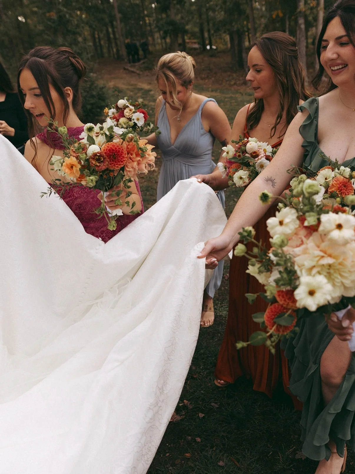 A group of women holding bouquets of flowers, helping to carry a wedding dress outdoors in a wooded area.