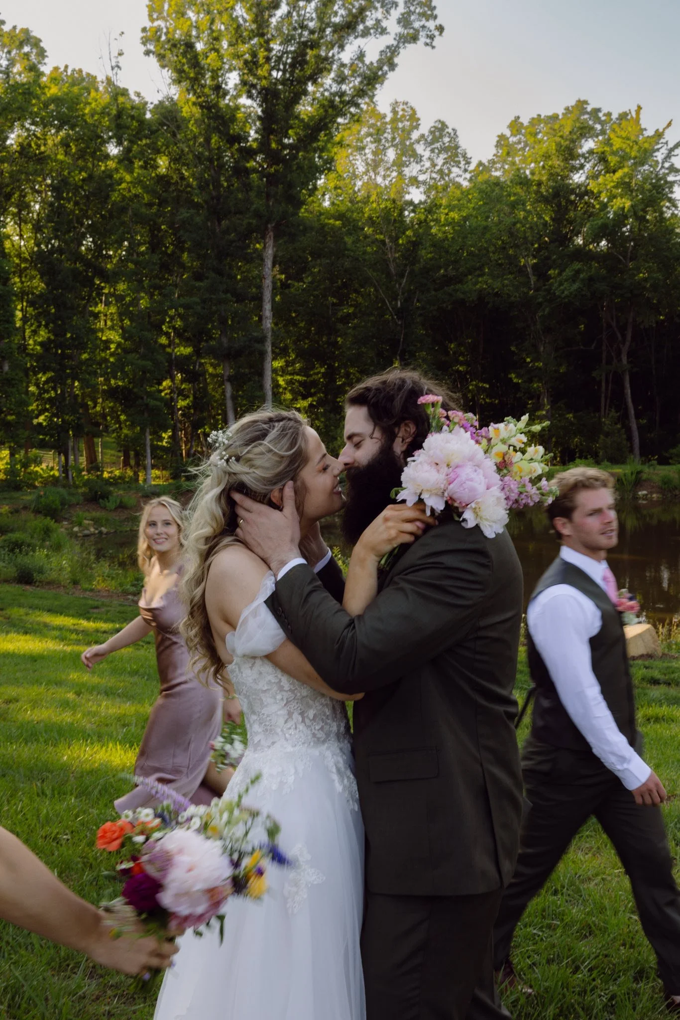 A bride and groom share a kiss during their outdoor wedding, holding a bouquet of pink and white flowers, with bridesmaids and groomsmen in the background on a grassy area near a pond, surrounded by trees.