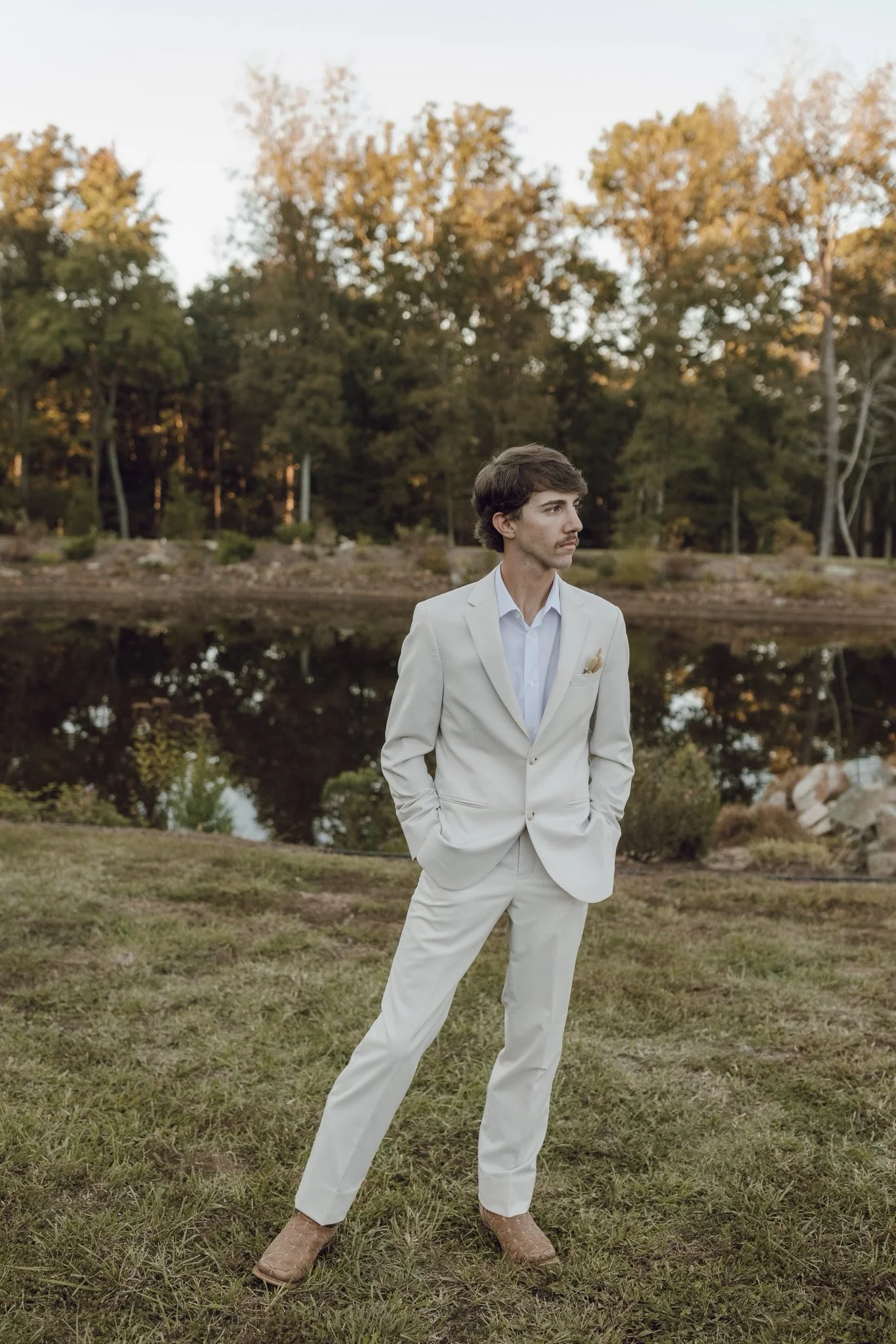 A man in a cream-colored suit stands outdoors near a body of water with trees in the background.