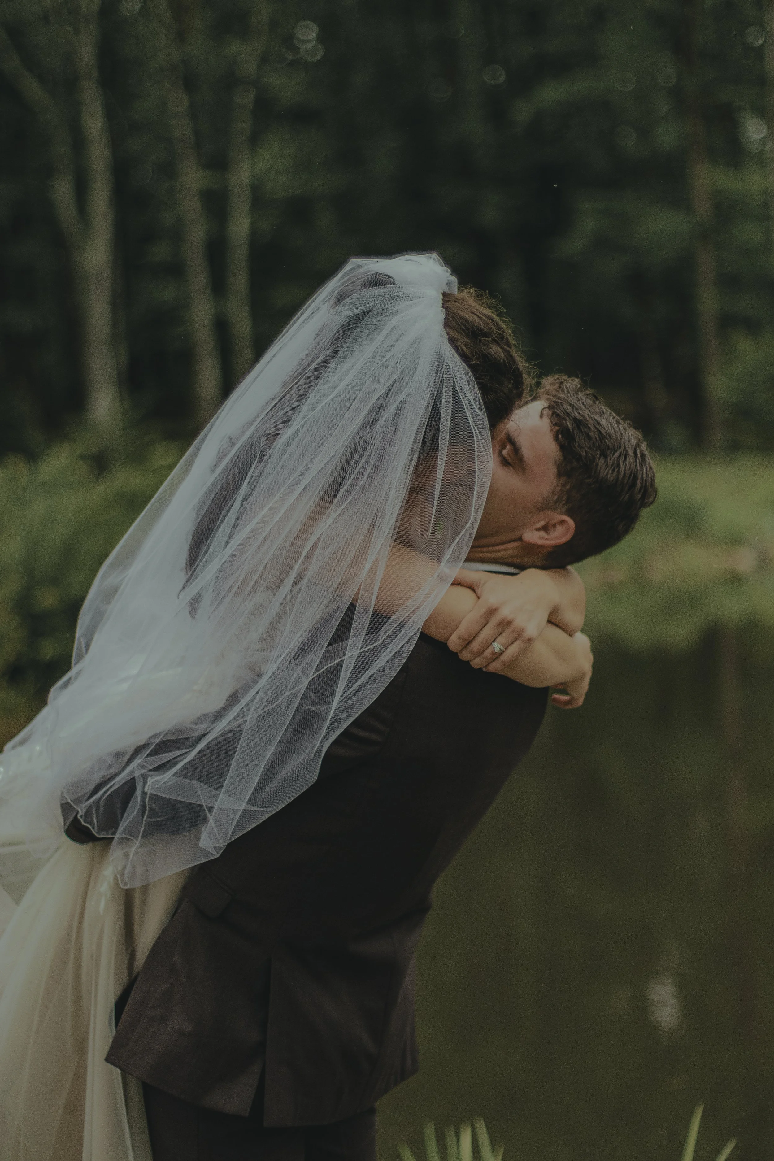A newlywed couple embracing outdoors near a body of water, with the woman wearing a veil and the man in a tuxedo, in a romantic moment surrounded by trees.