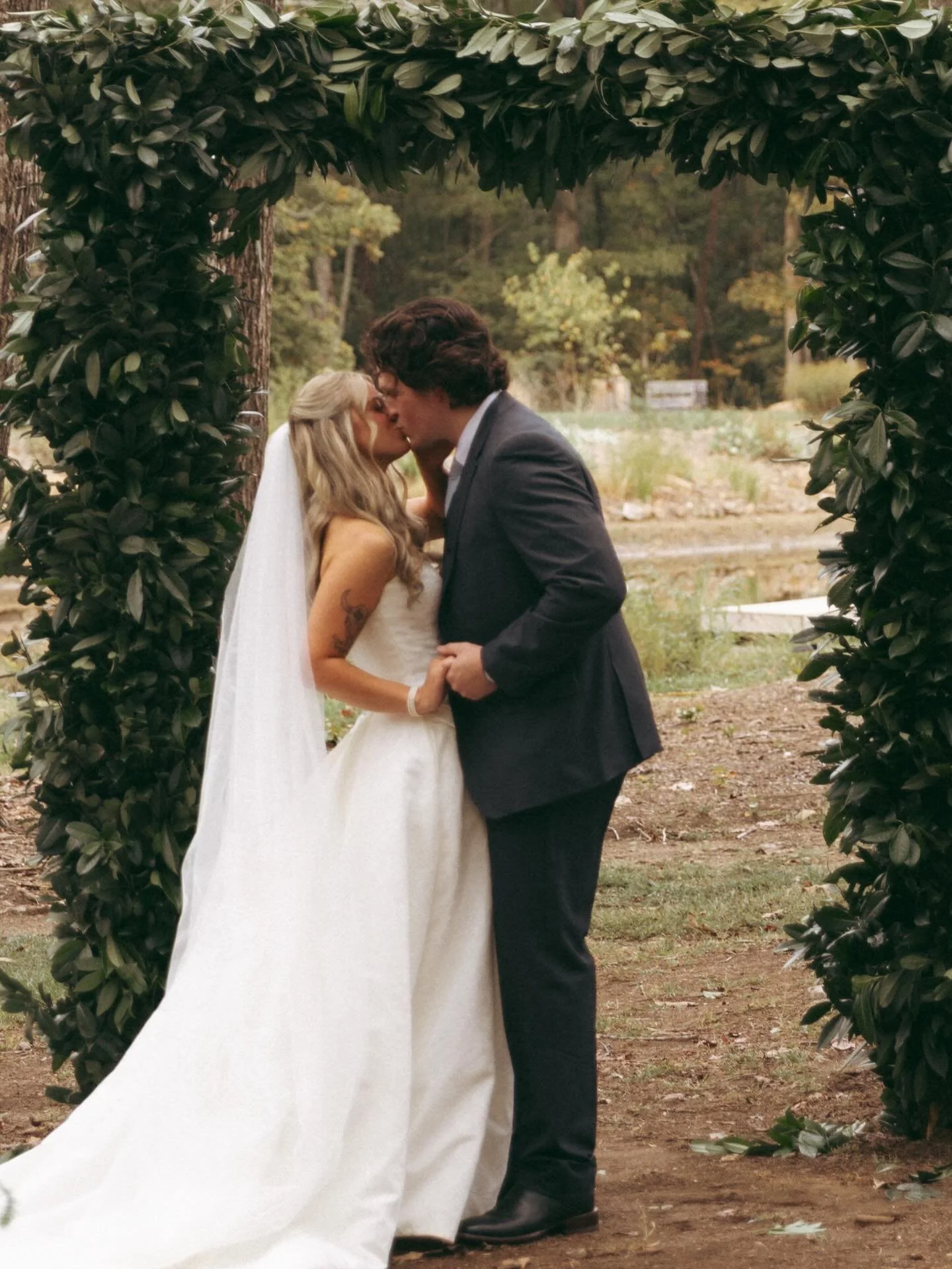 A bride and groom share a kiss under a green arch at an outdoor wedding ceremony, with trees and natural scenery in the background.