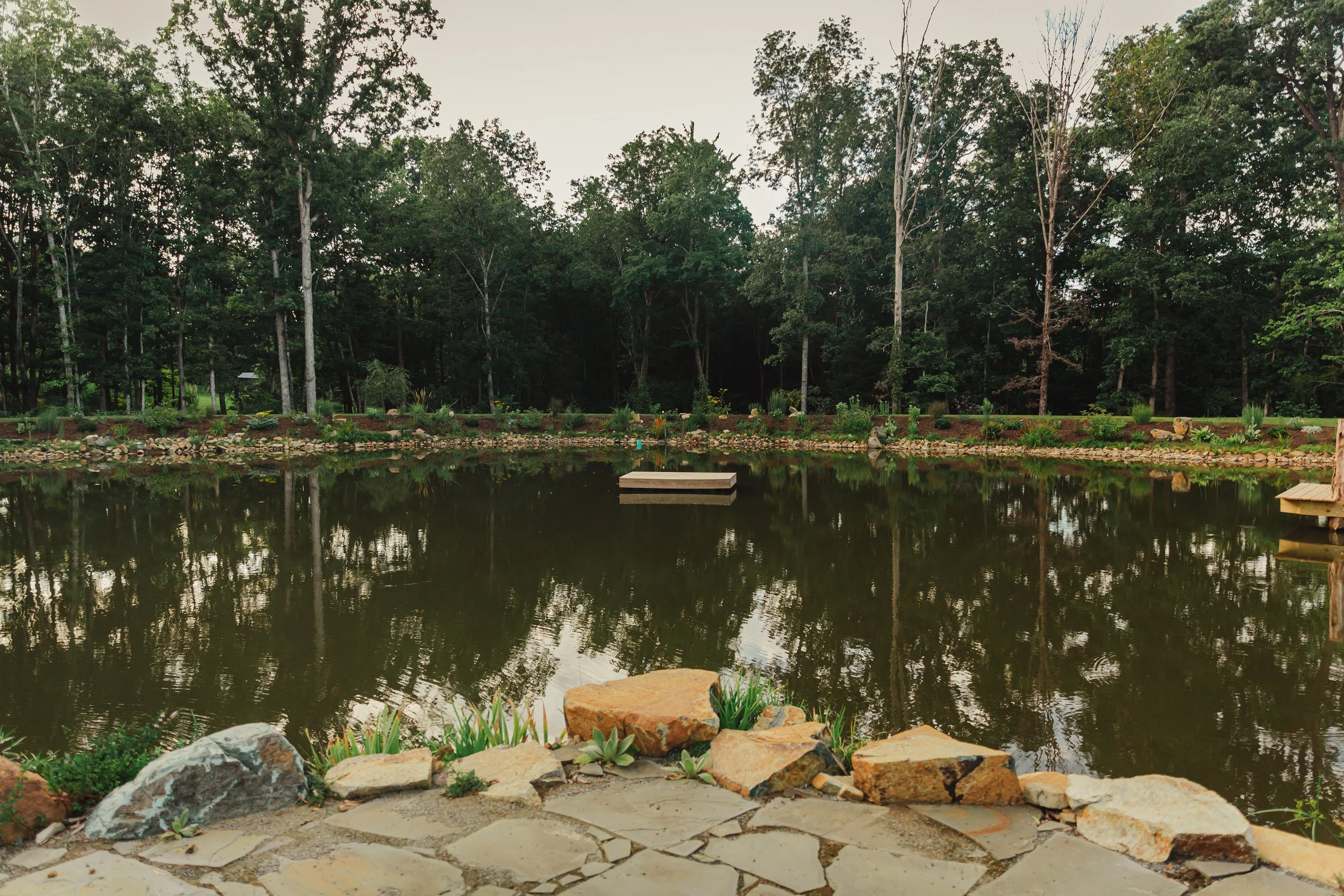 A peaceful pond bordered by rocks with surrounding lush green trees and a cloudy sky overhead.