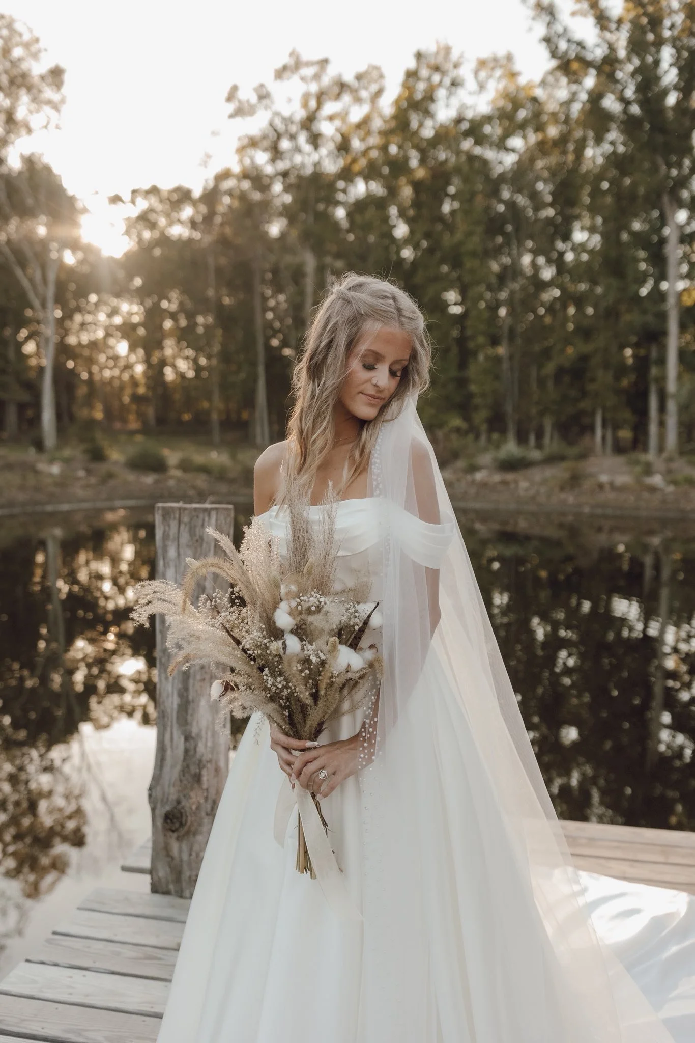 A bride in a white wedding dress holding a bouquet of dried flowers, standing on a wooden dock near a pond, with trees and sunlight in the background.