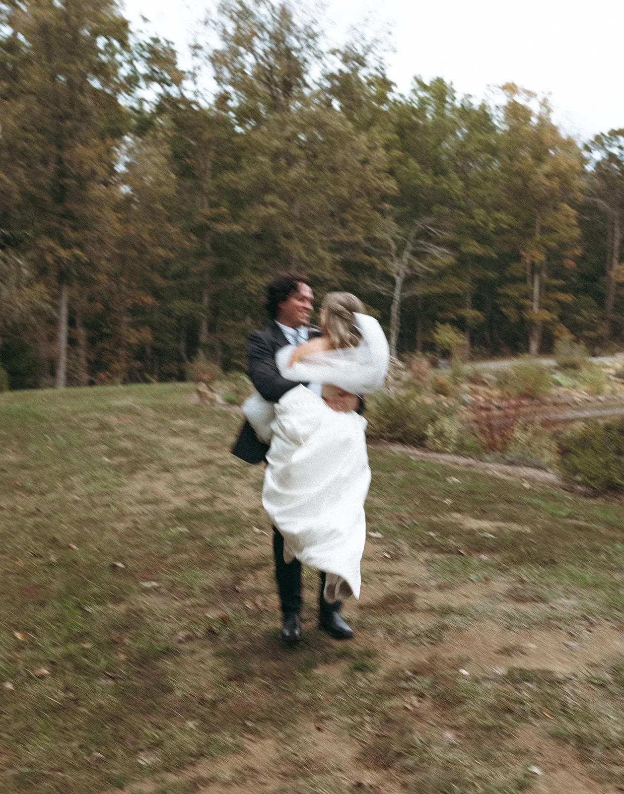 A person in formal wedding attire, carrying a bride dressed in white, running outdoors on a grassy area with trees in the background.