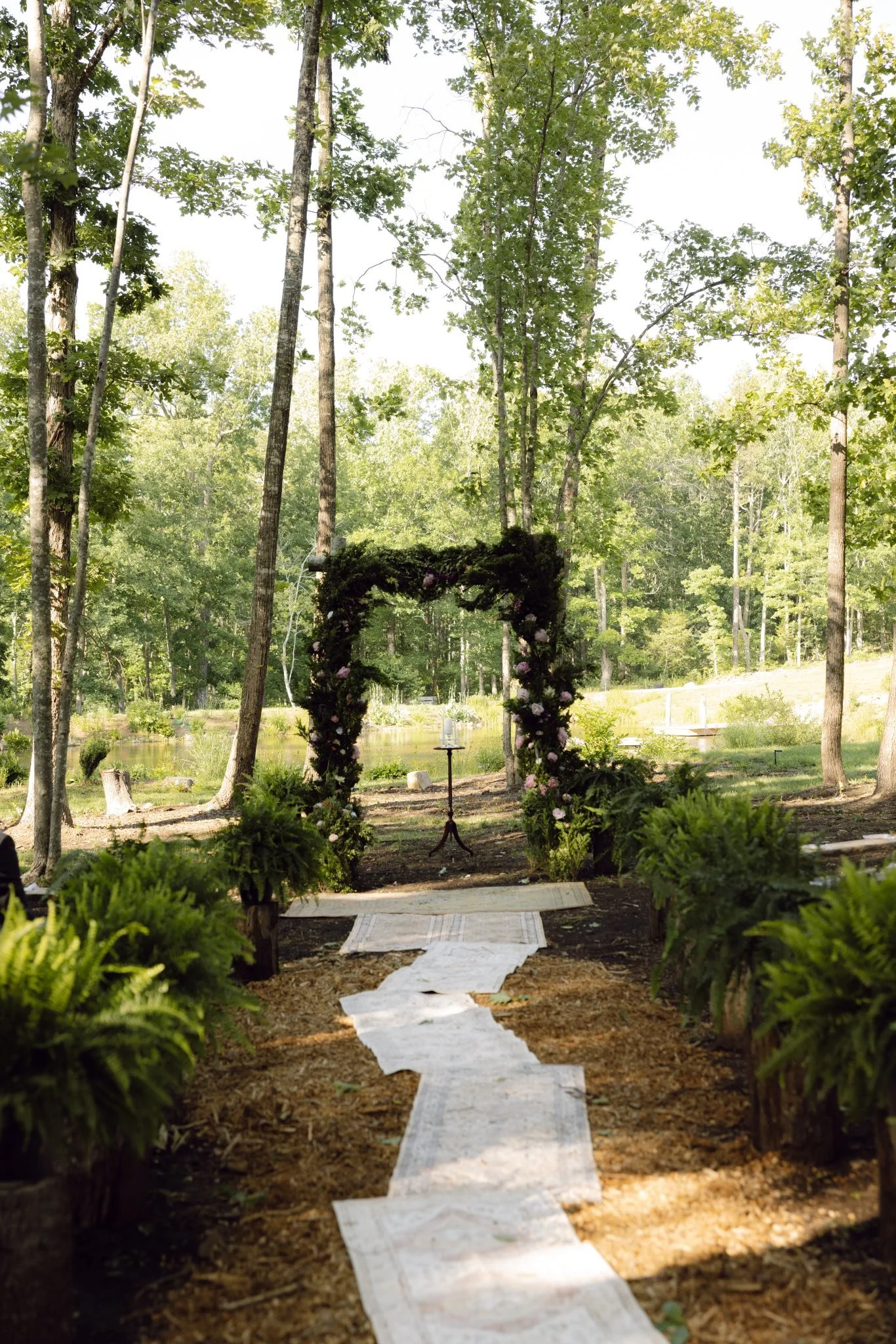 Outdoor wedding ceremony setup with an arch decorated with greenery and flowers, a pathway lined with rugs, and tall trees surrounding the area.