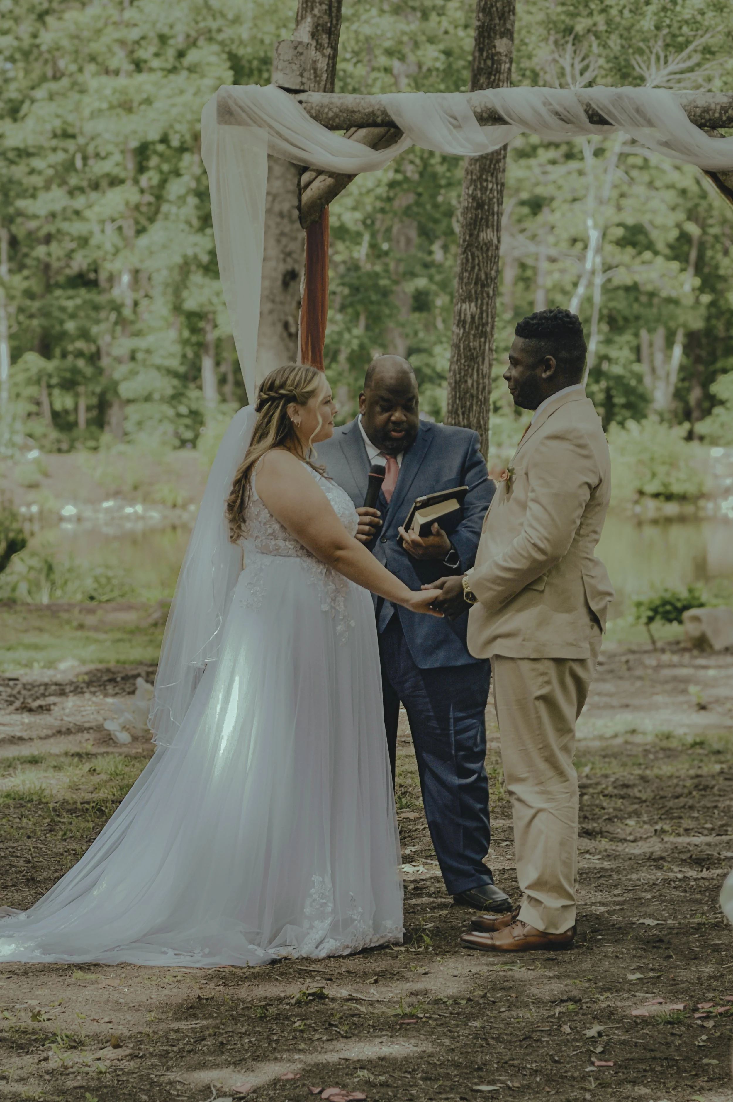 A bride and groom holding hands and exchanging vows during an outdoor wedding ceremony in a wooded area, with a officiant standing between them.