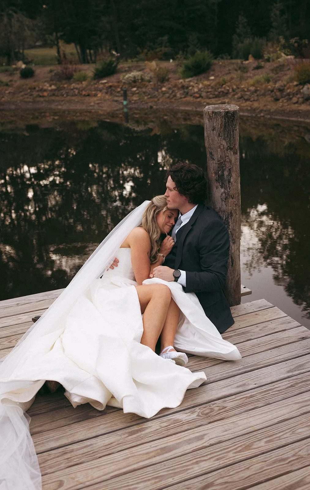 A bride and groom sitting closely on a wooden dock by a calm river, embracing each other with eyes closed, surrounded by nature.