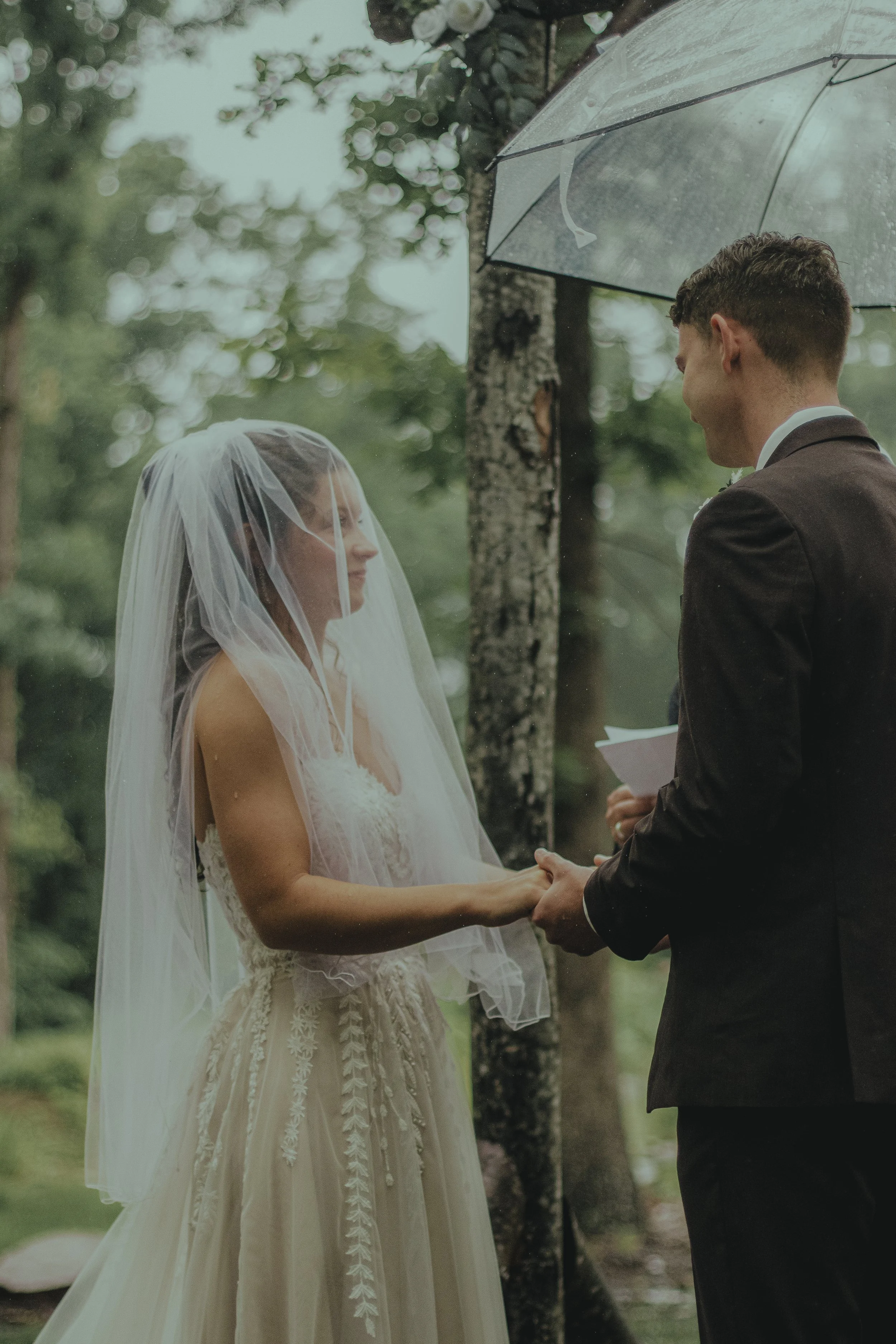 A bride and groom holding hands during an outdoor wedding ceremony in the rain, with the officiant behind them, under a clear umbrella, in a lush green forest setting.