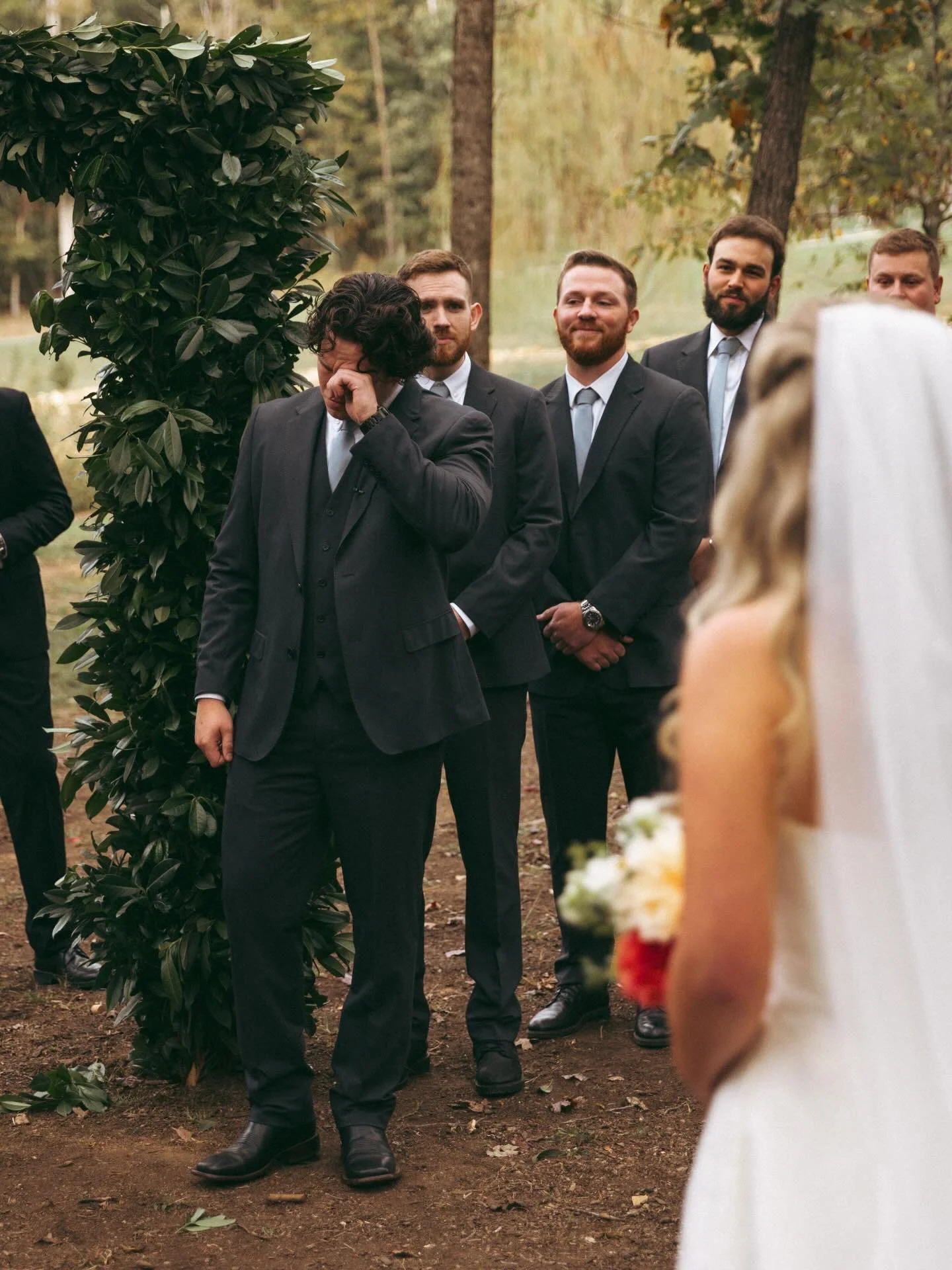 Man in suit wiping away tears or emotional moment during outdoor wedding ceremony with friends or groomsmen in formal suits watching in the woods.