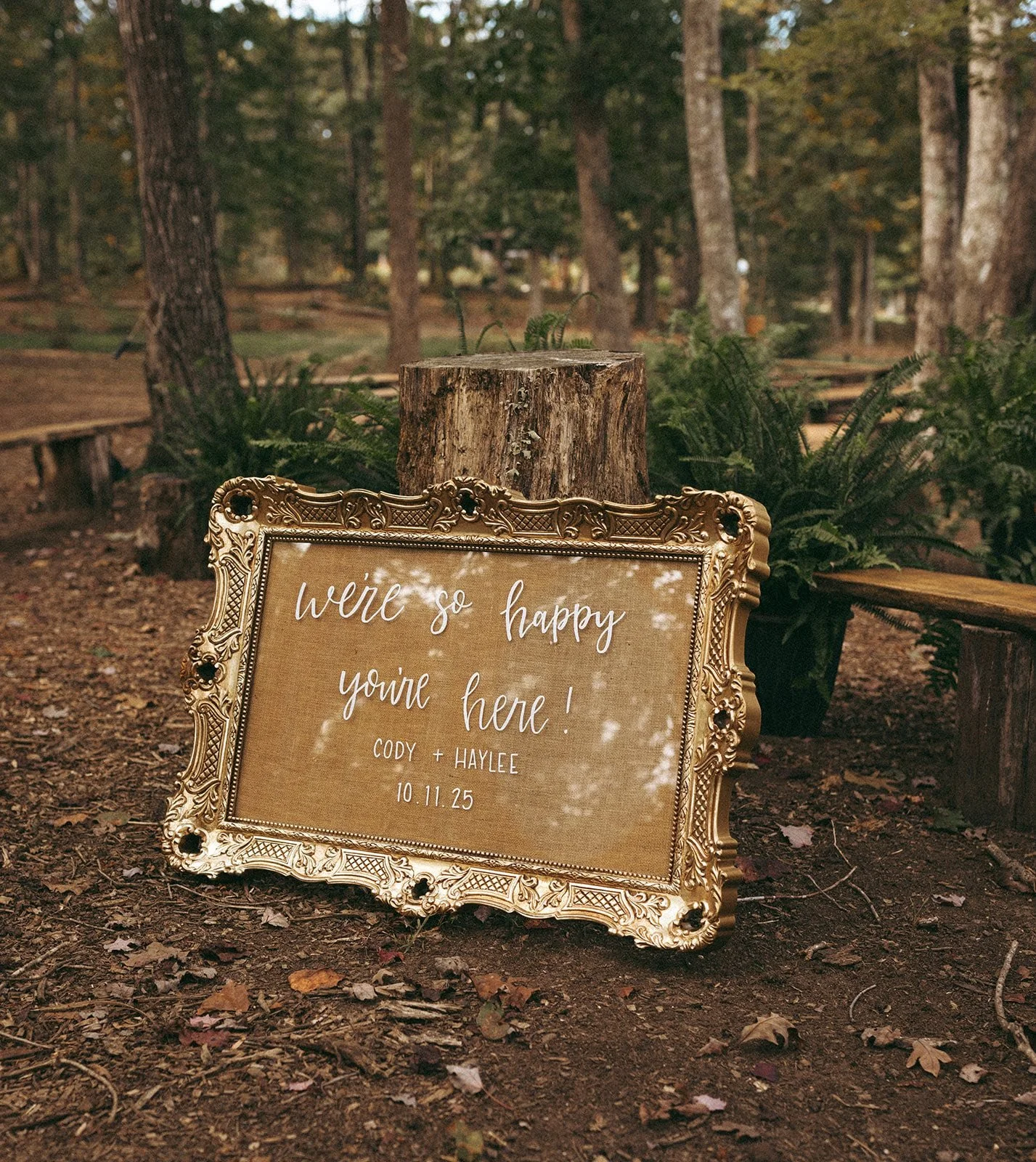 A decorative sign with gold ornate frame on the ground in a wooded outdoor area, reading 'we're so happy you're here! CODY + HAYLEE 10.11.25', surrounded by ferns and trees.