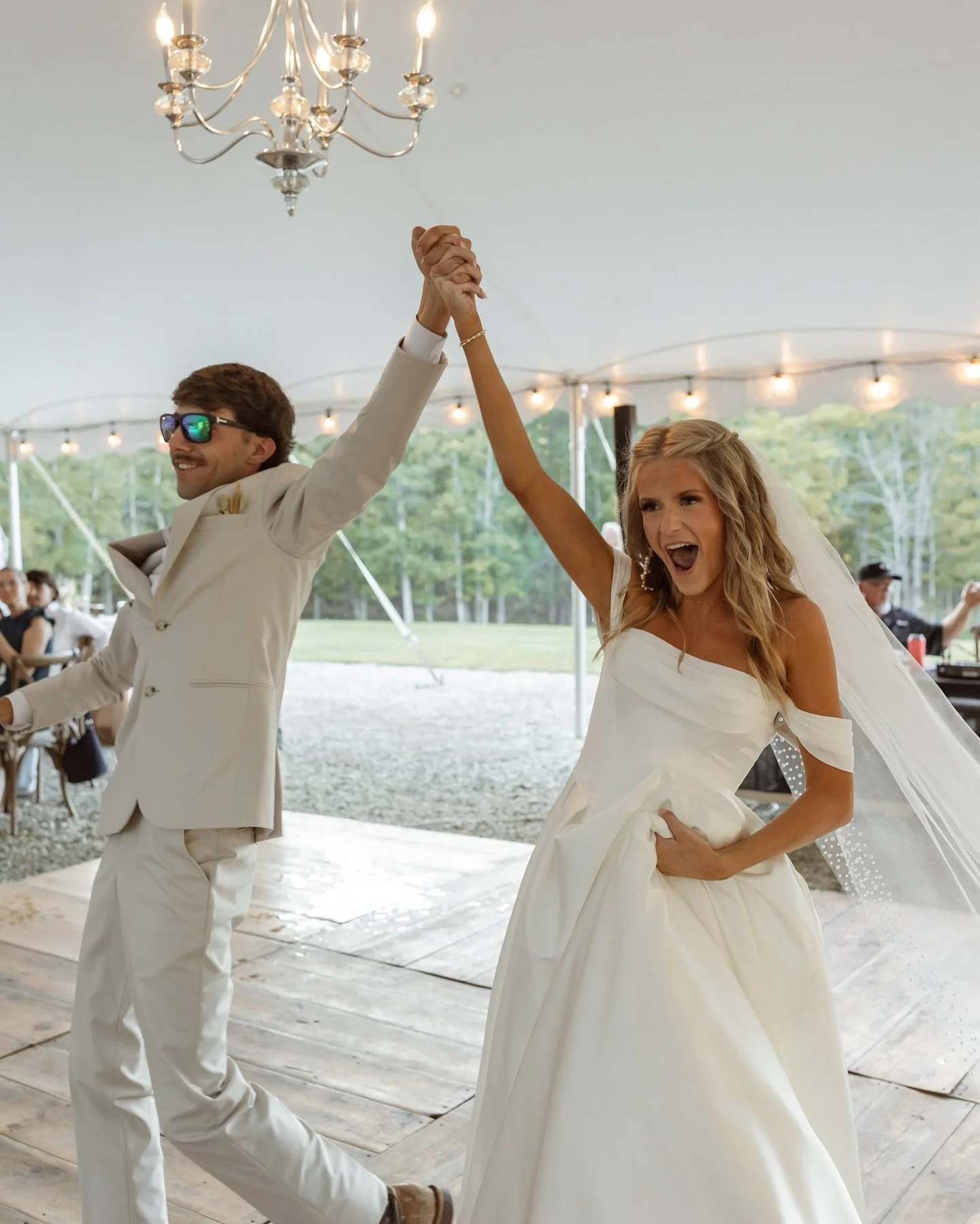 Bride and groom dancing and holding hands at their wedding reception under a tent with string lights and a chandelier.