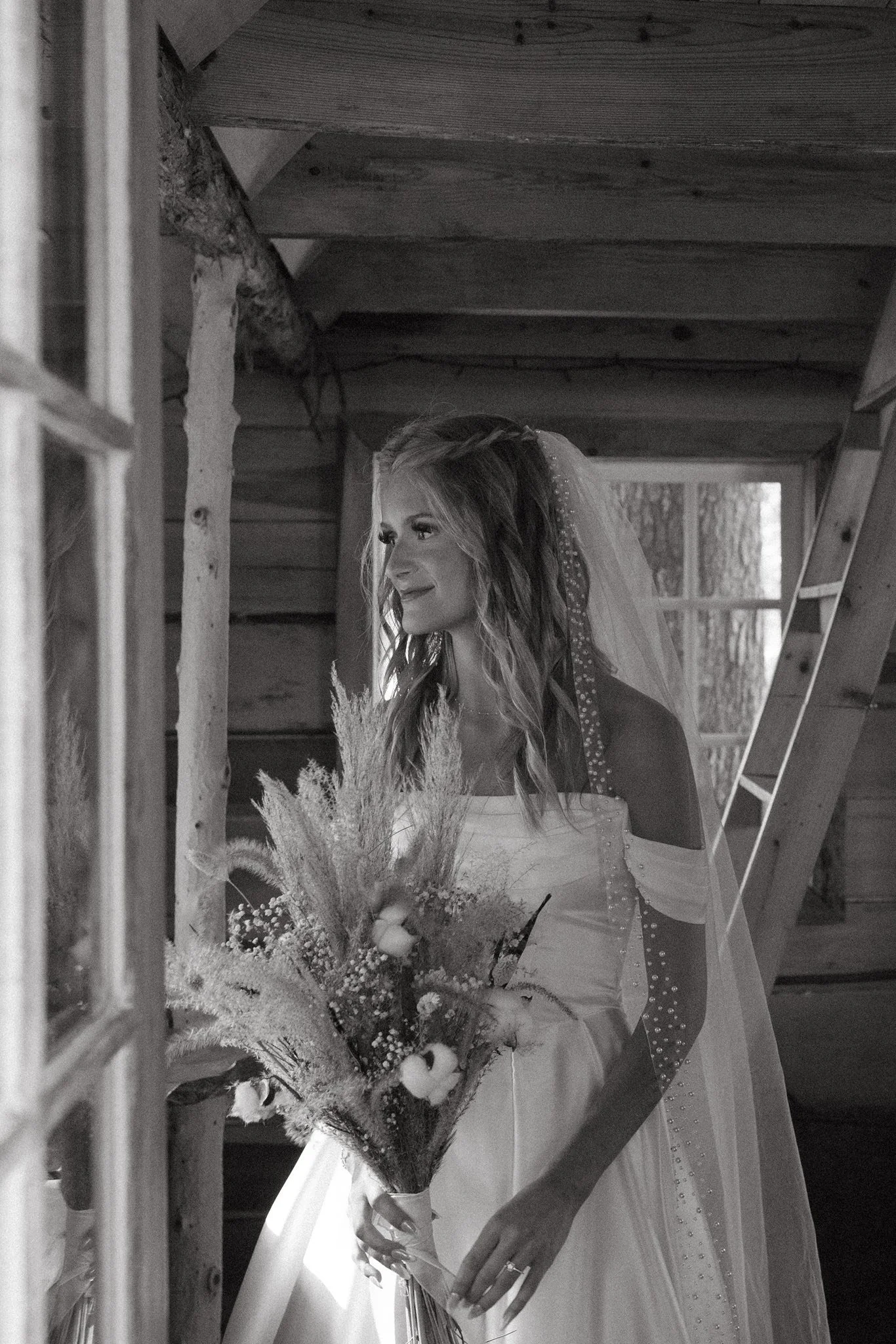 Black and white photo of a smiling bride holding a bouquet of flowers, inside a wooden cabin.