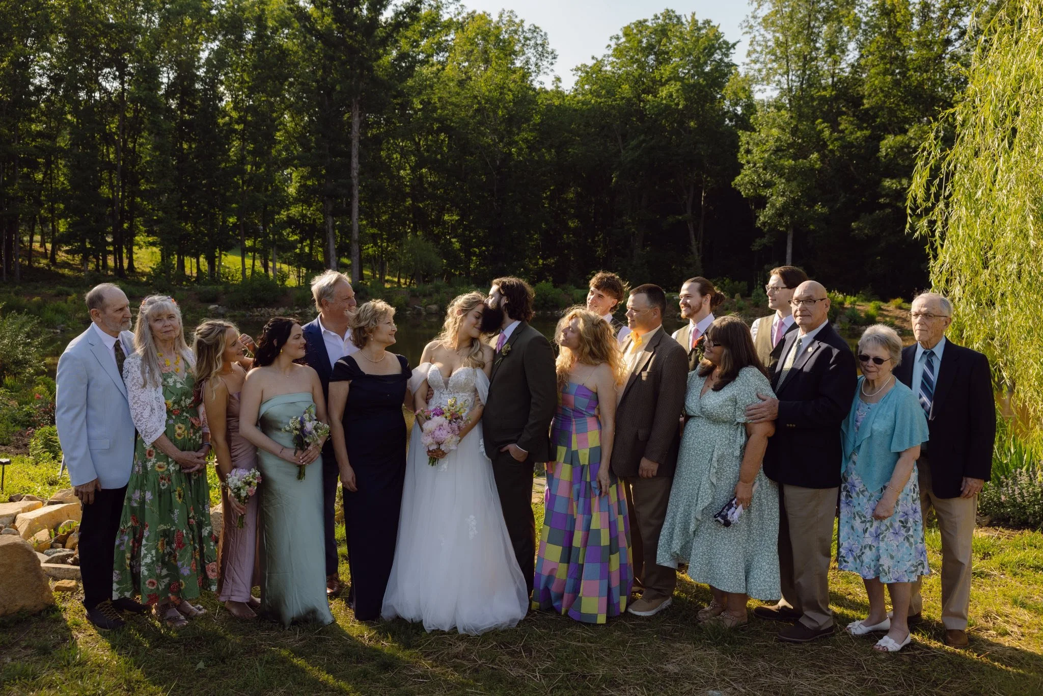 A group of people at a wedding outside near a pond, with trees in the background. The bride and groom are in the center, surrounded by family and friends.