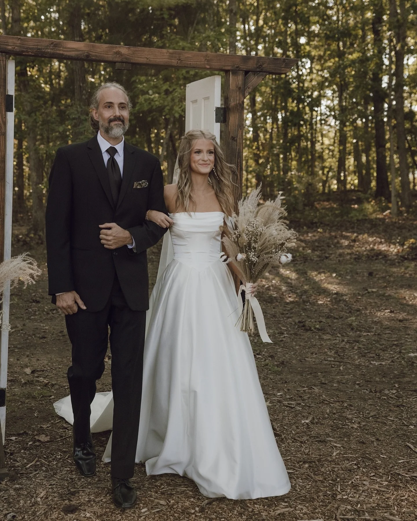 A bride in a white wedding gown holding a bouquet of dried flowers stands next to a man in a black suit during an outdoor wedding ceremony in a wooded area, with a wooden arch and natural foliage in the background.