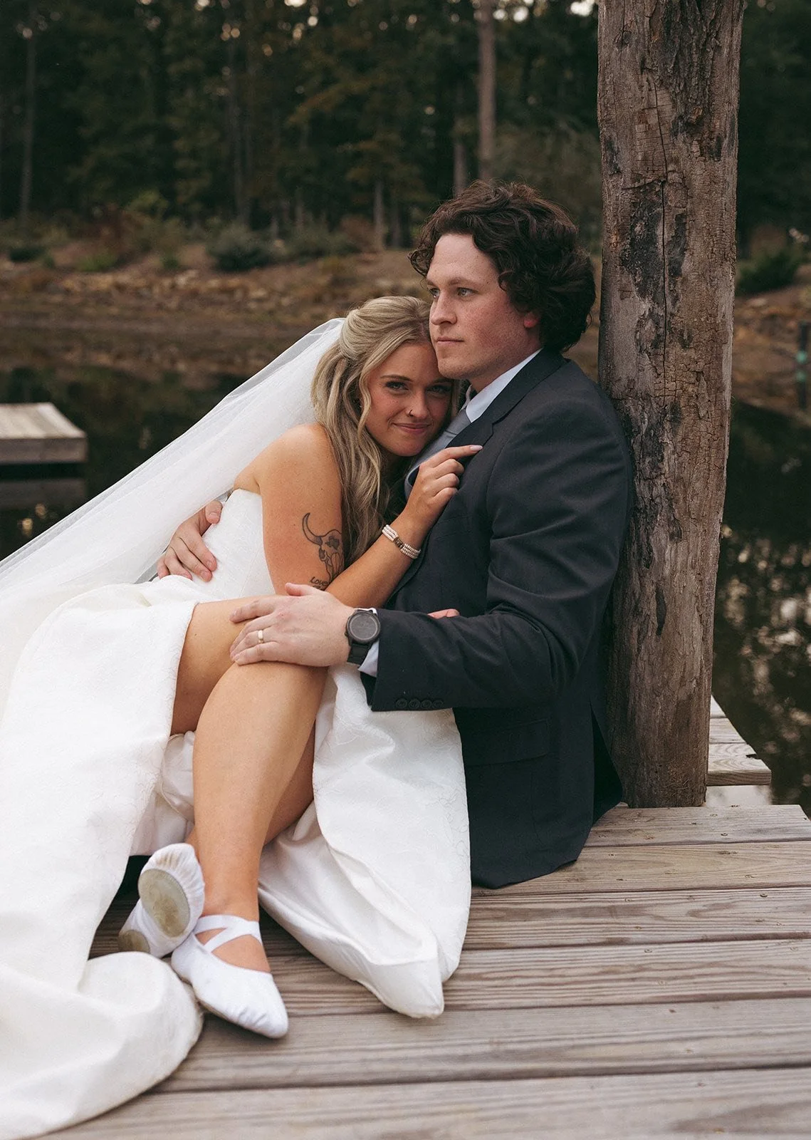 A newlywed couple sitting on a wooden dock by a lake, with the bride resting her head on the groom's shoulder, outdoors with trees in the background.