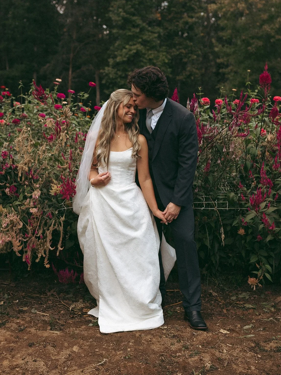 A bride and groom standing close together outdoors in front of pink and purple flowers, with the groom kissing the bride on her forehead.