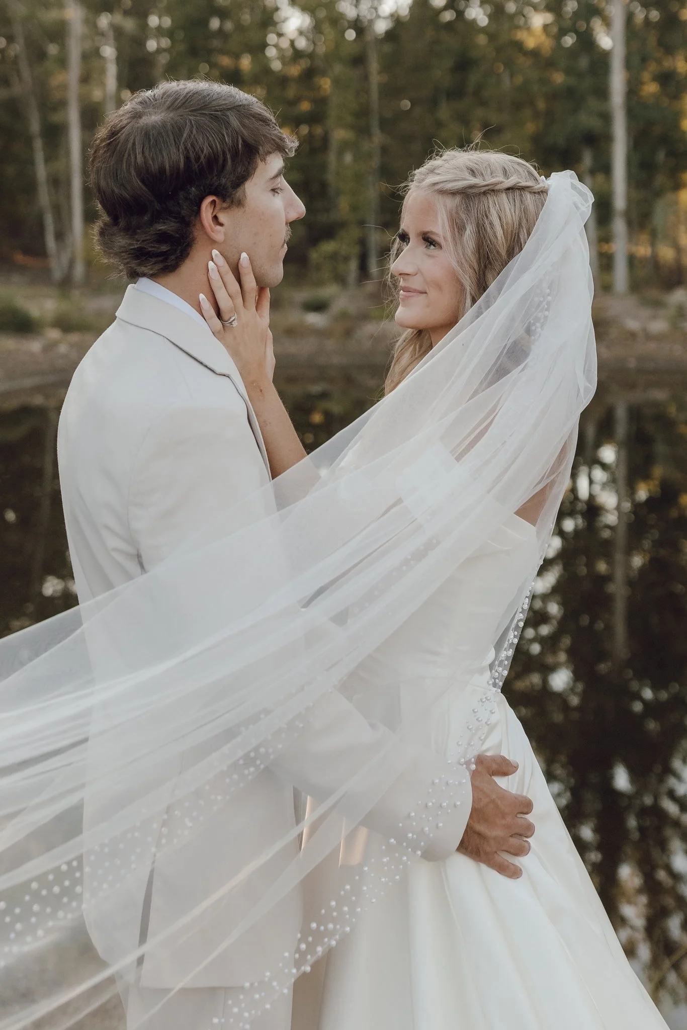 A bride and groom standing close by a lake in a forest. They look at each other affectionately, with the bride holding the groom's face. The bride is wearing a white wedding gown and veil, and the groom is in a white suit. The setting appears to be d