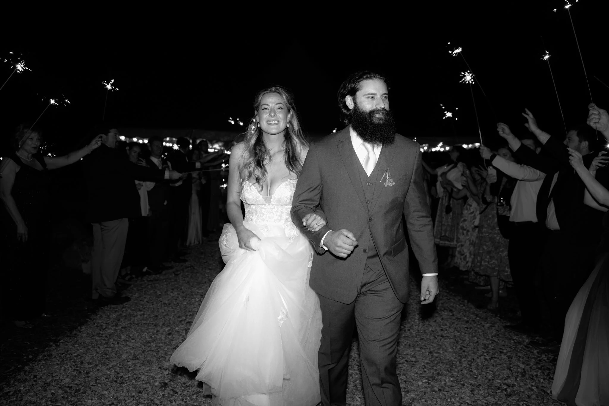 A black-and-white photo of a newlywed couple walking together, surrounded by guests holding sparklers at night. The bride is wearing a white wedding dress, and the groom is dressed in a suit with a beard.