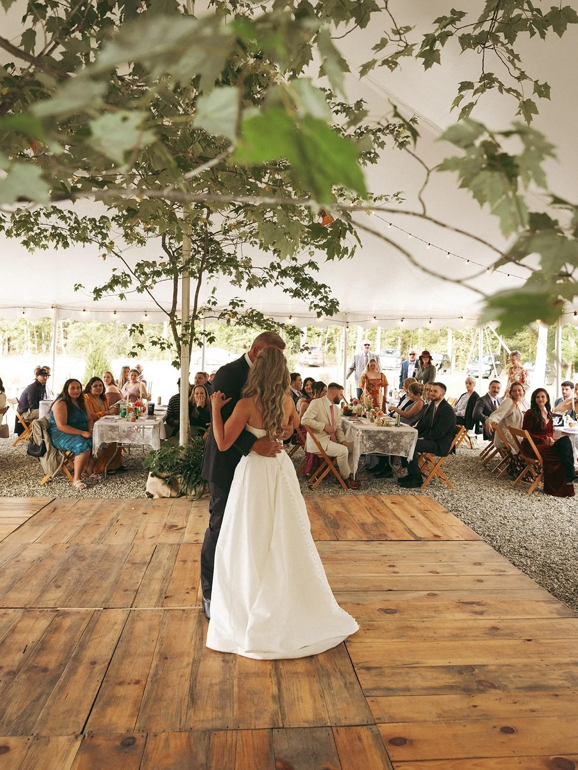 A bride and groom dancing in the center of an outdoor wedding reception under a large tent, with guests seated at tables in the background.