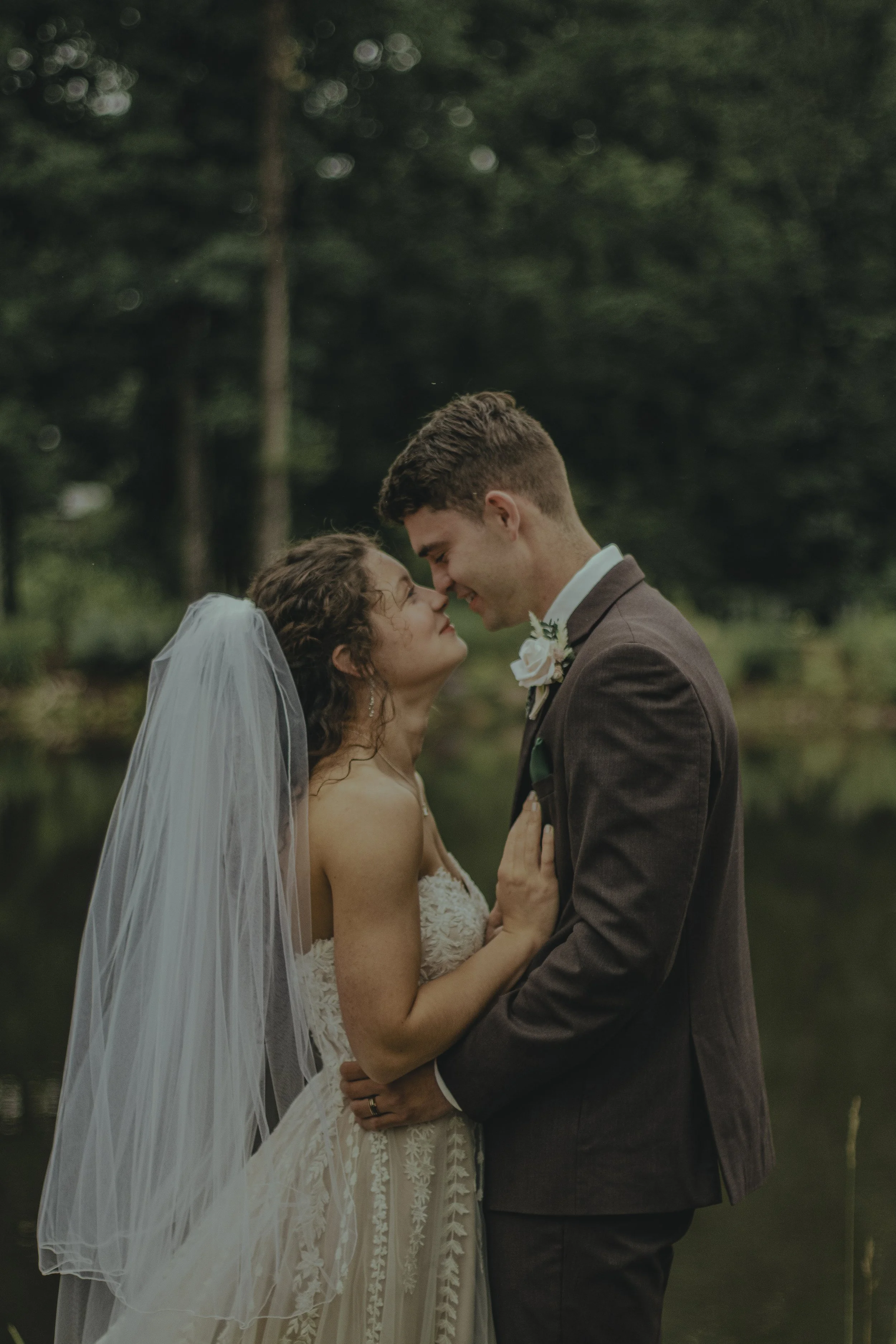 A bride and groom share an intimate moment, standing close with their foreheads touching, outdoors near a body of water with a wooded background.