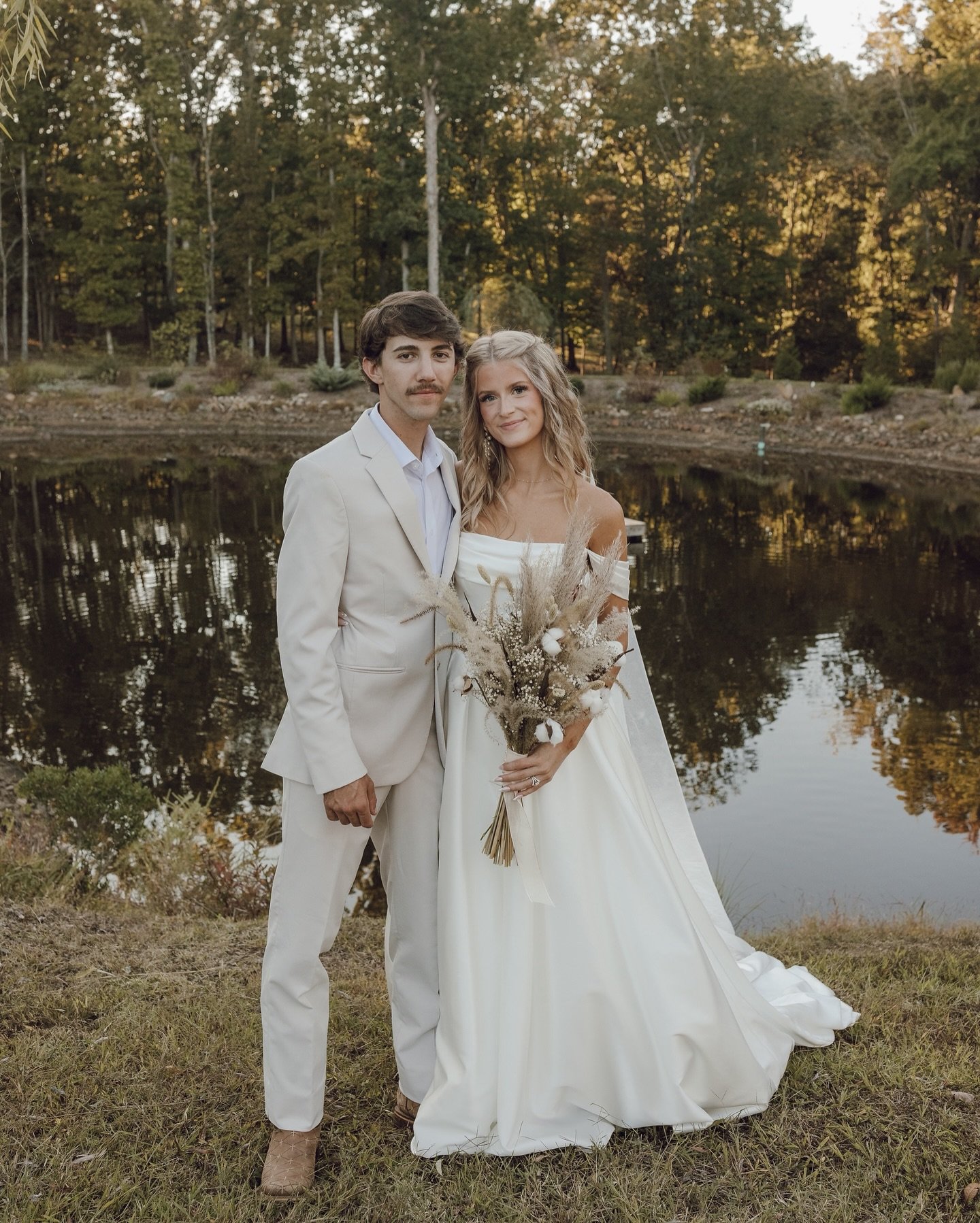 A couple dressed in wedding attire standing by a pond surrounded by trees at sunset, with the bride holding a bouquet of dried flowers.