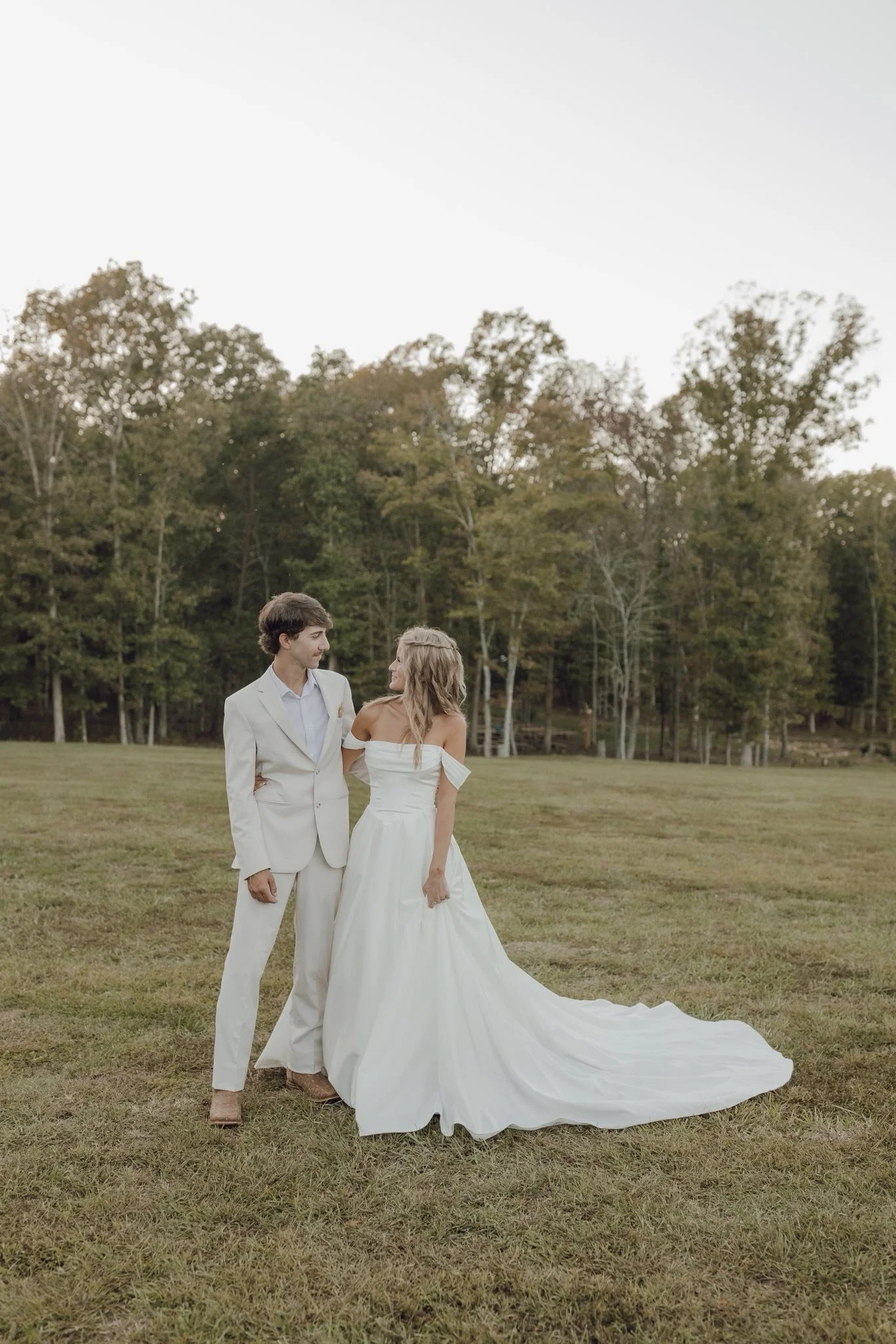 A bride and groom standing in a grassy field with trees in the background, dressed in wedding attire, gazing at each other.